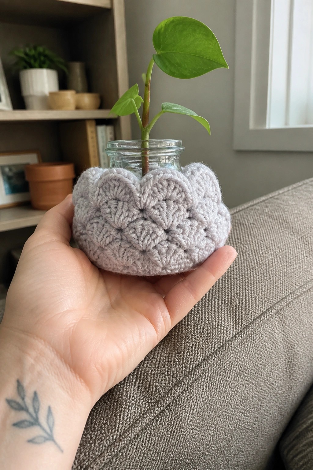 Hand with leaf tattoo holds gray crocheted cozy around glass jar with plant
