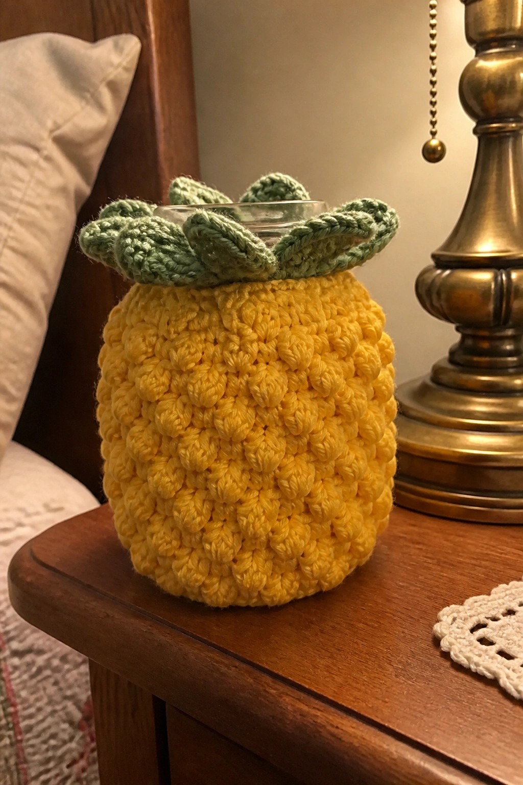 Yellow crocheted pineapple jar cozy with green leaves on wooden table by lamp.