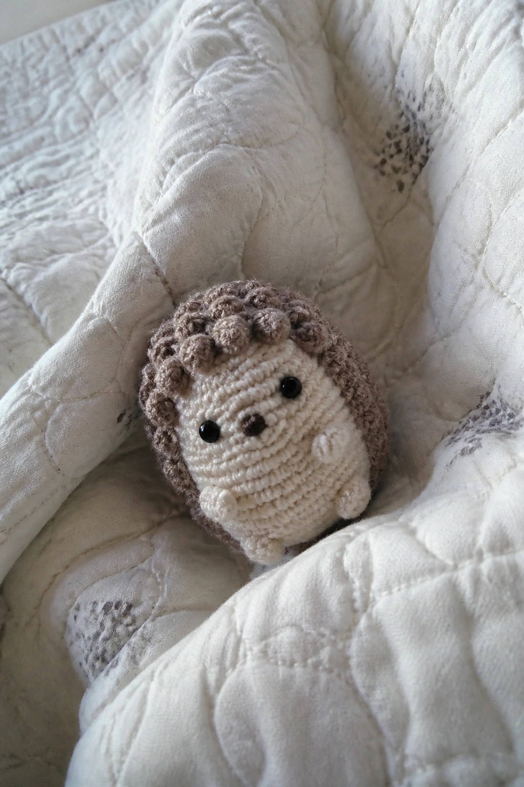 Tiny crocheted hedgehog with brown spikes on white quilted blanket.