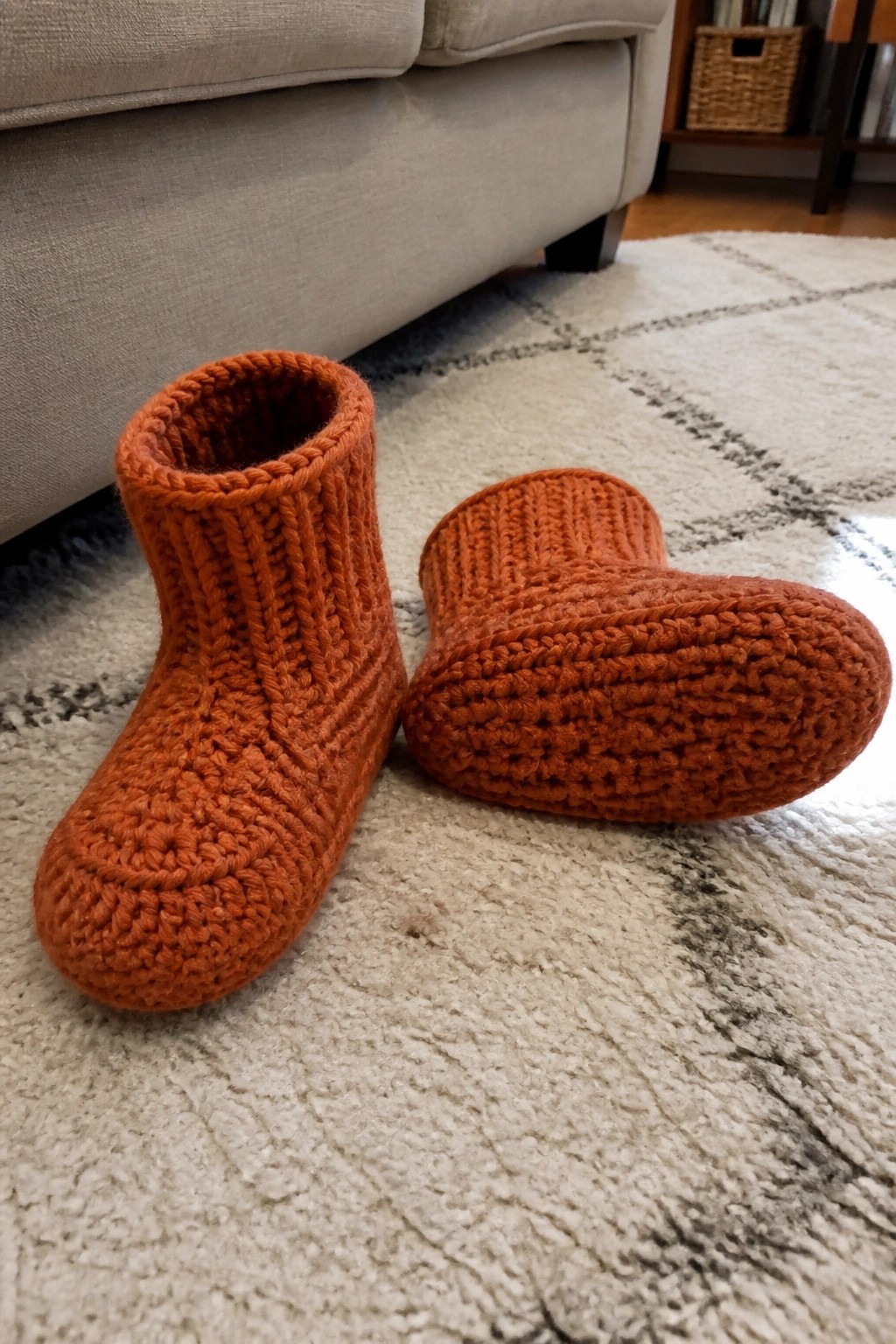 A pair of orange crocheted slippers on a rug, one upright and one on its side.