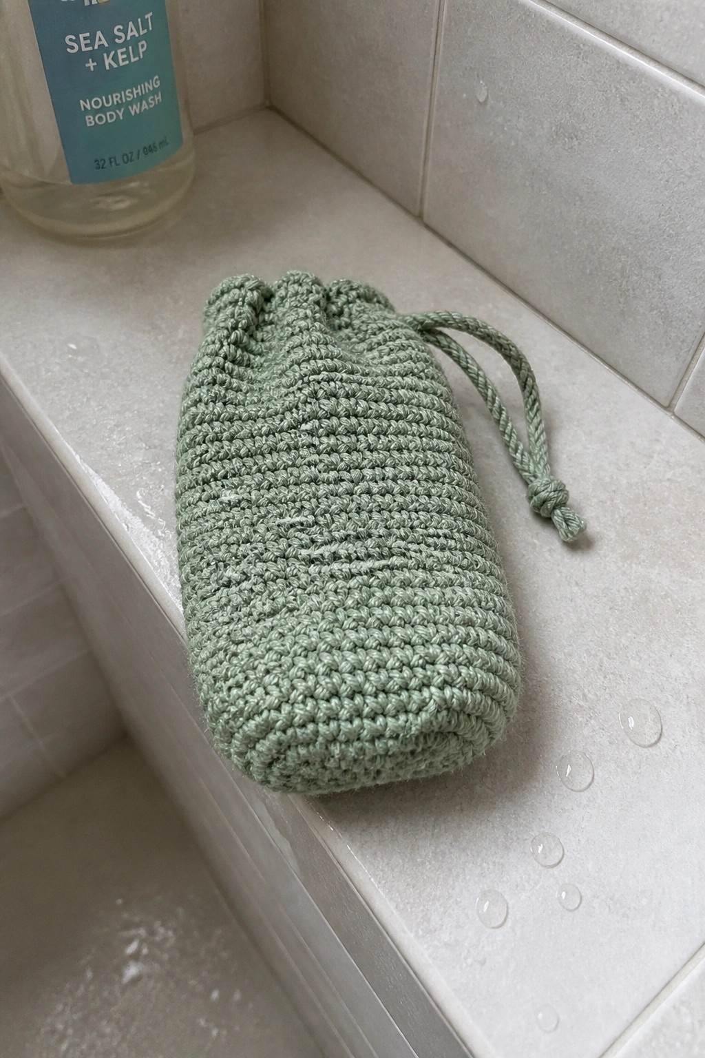 Sage green crocheted drawstring pouch on wet shower ledge beside body wash bottle