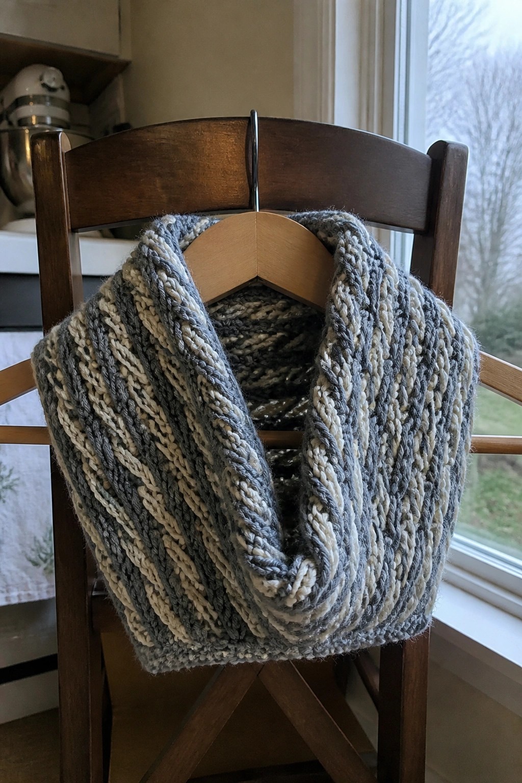 Chunky gray-and-white striped knit cowl on wooden hanger over chair.
