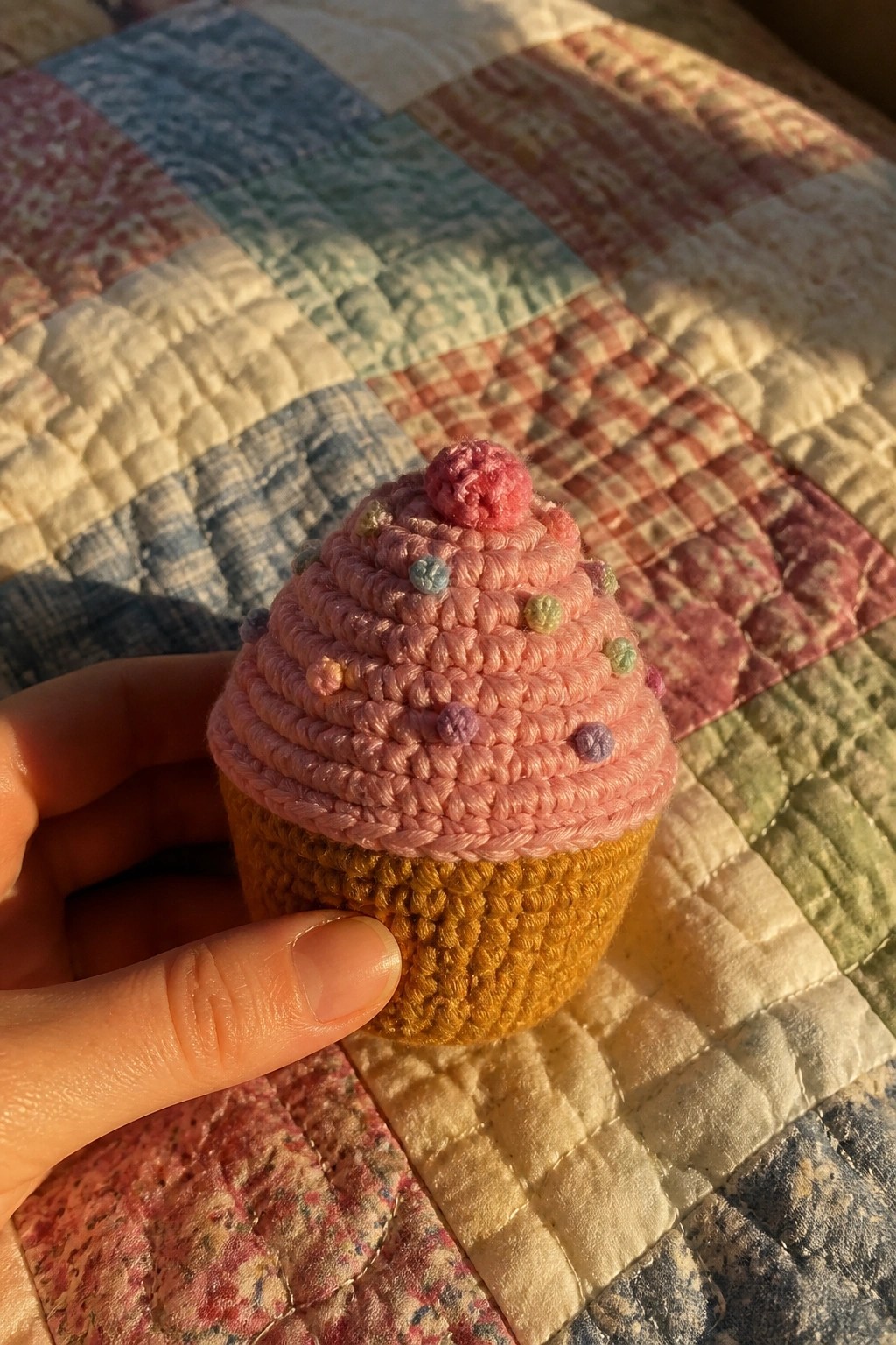 Hand holding pink crocheted cupcake with colorful sprinkles atop patchwork quilt in sunlight