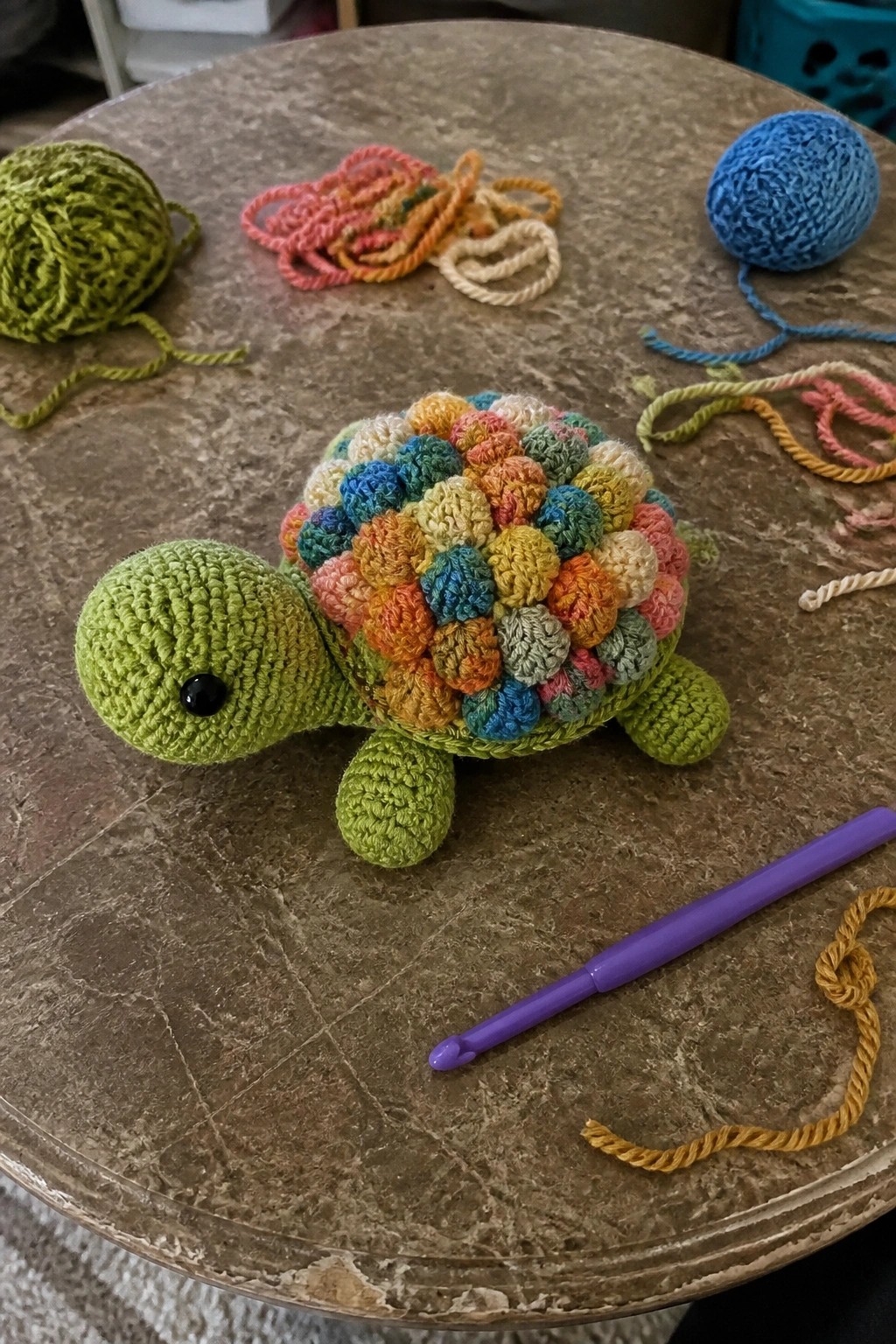 Crocheted green turtle with multicolored bobble shell on a table beside a purple hook and yarn balls.