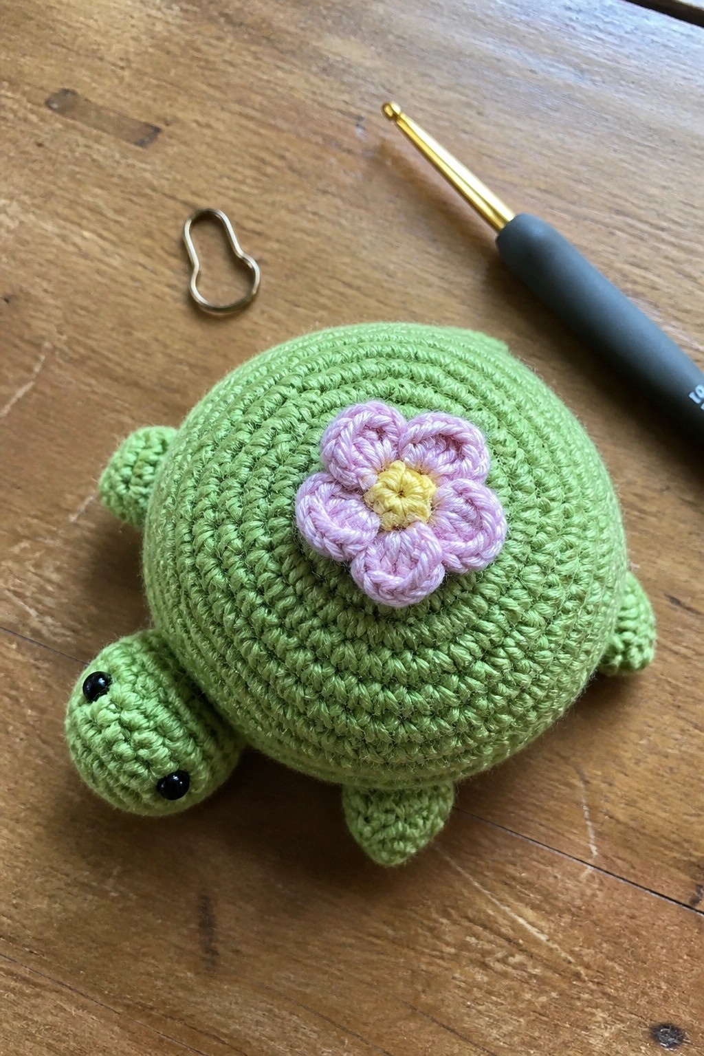 Green crocheted turtle with pink flower beside hook and stitch marker on wood