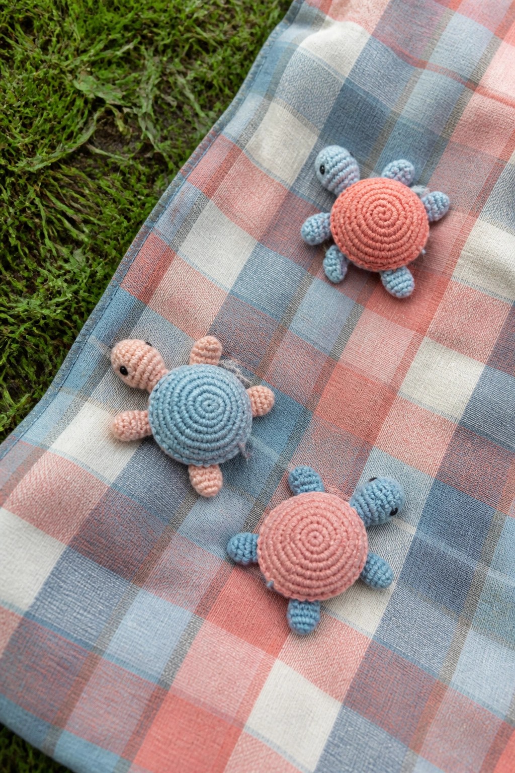 Three crocheted turtles with pink and blue shells on plaid blanket over grass