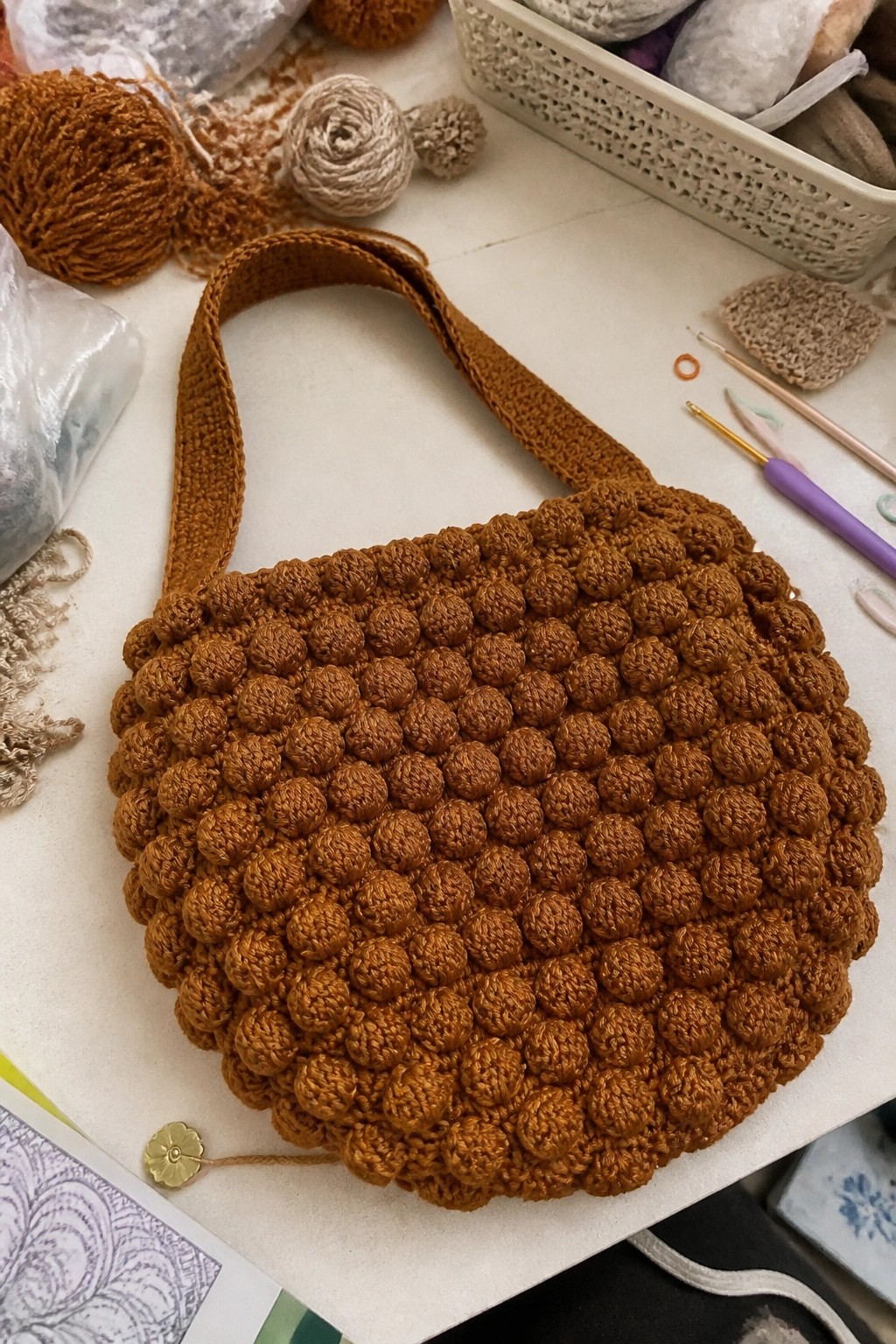 Brown crocheted bobble-stitch bag with strap amid yarn balls and hooks