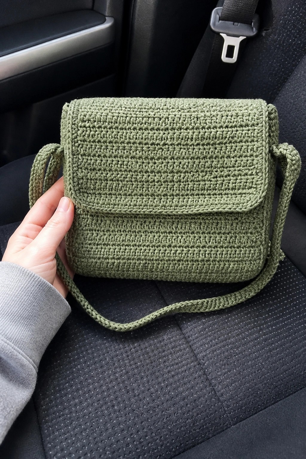 Hand holding green crocheted crossbody bag on black car seat with seatbelt
