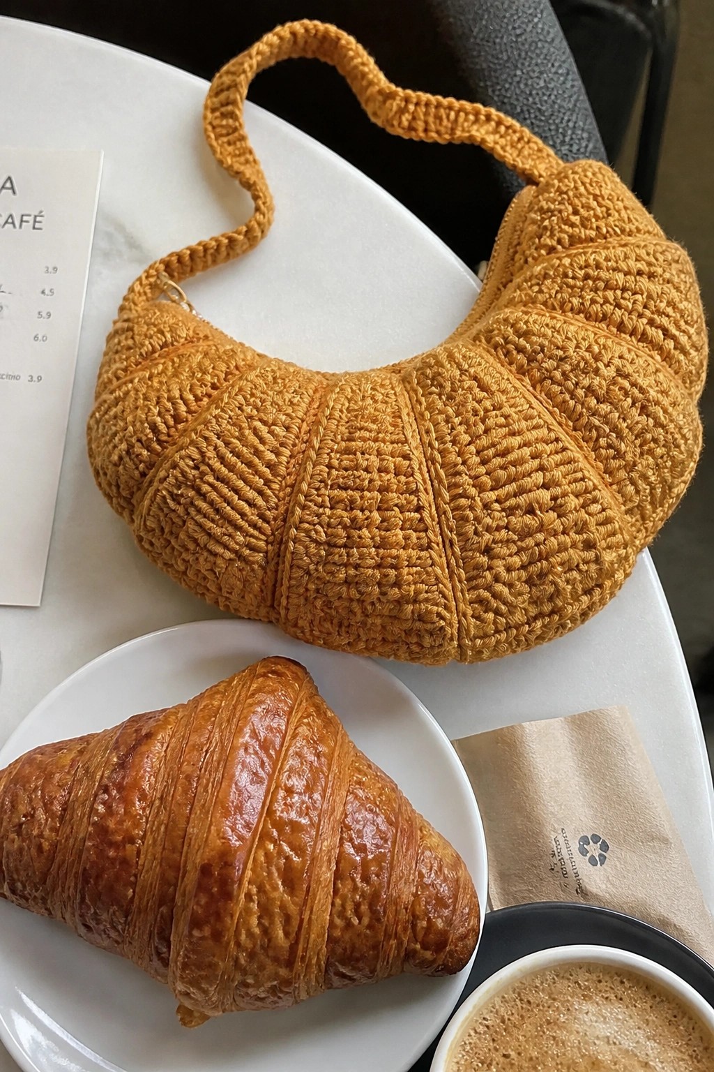 Crocheted orange croissant bag beside real pastry and coffee on cafe table