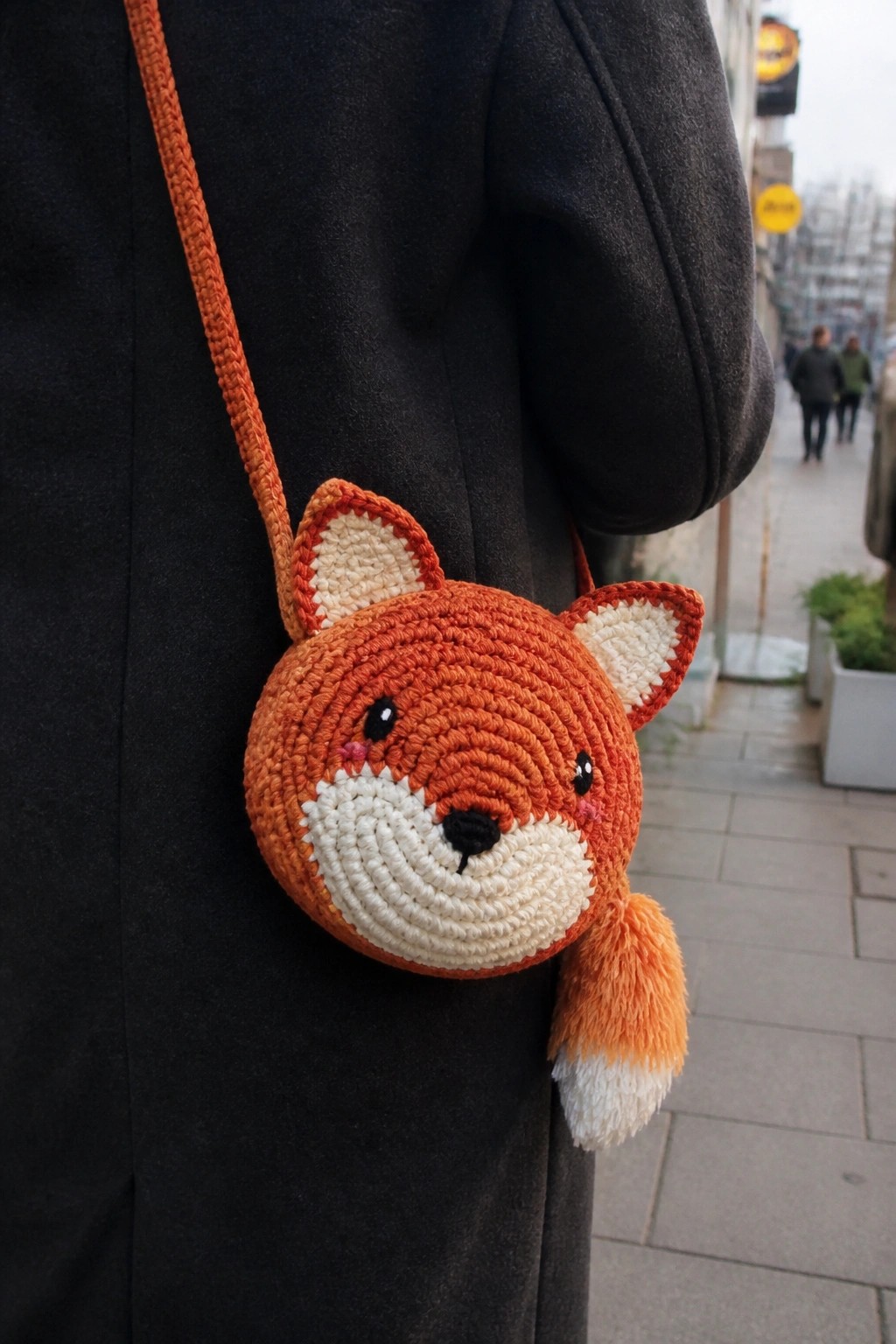 A person in a dark coat wearing an orange crocheted fox face shoulder bag with white muzzle, ears, and a small tail.