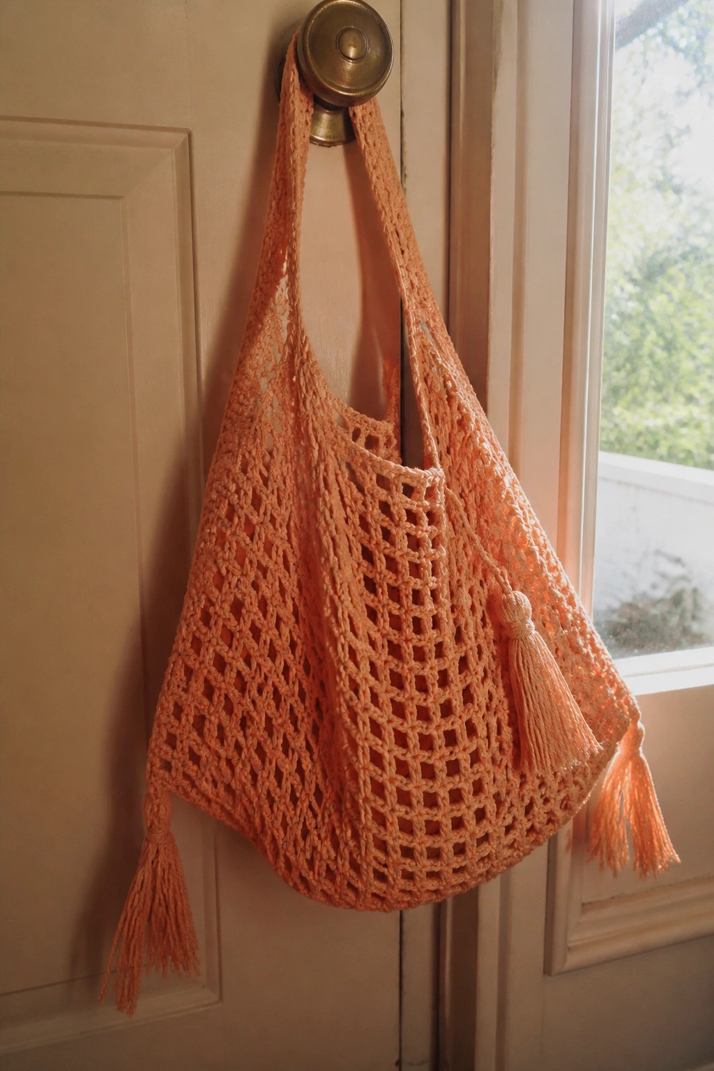 Peach crocheted net market bag with tassels hanging from brass doorknob