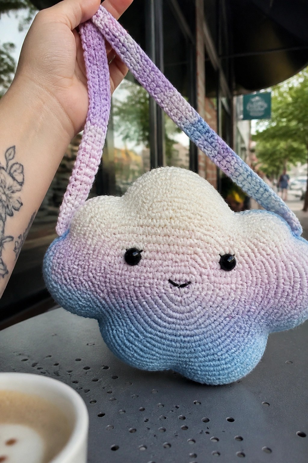 Hand holding pastel crocheted cloud bag with smiling face over coffee cup