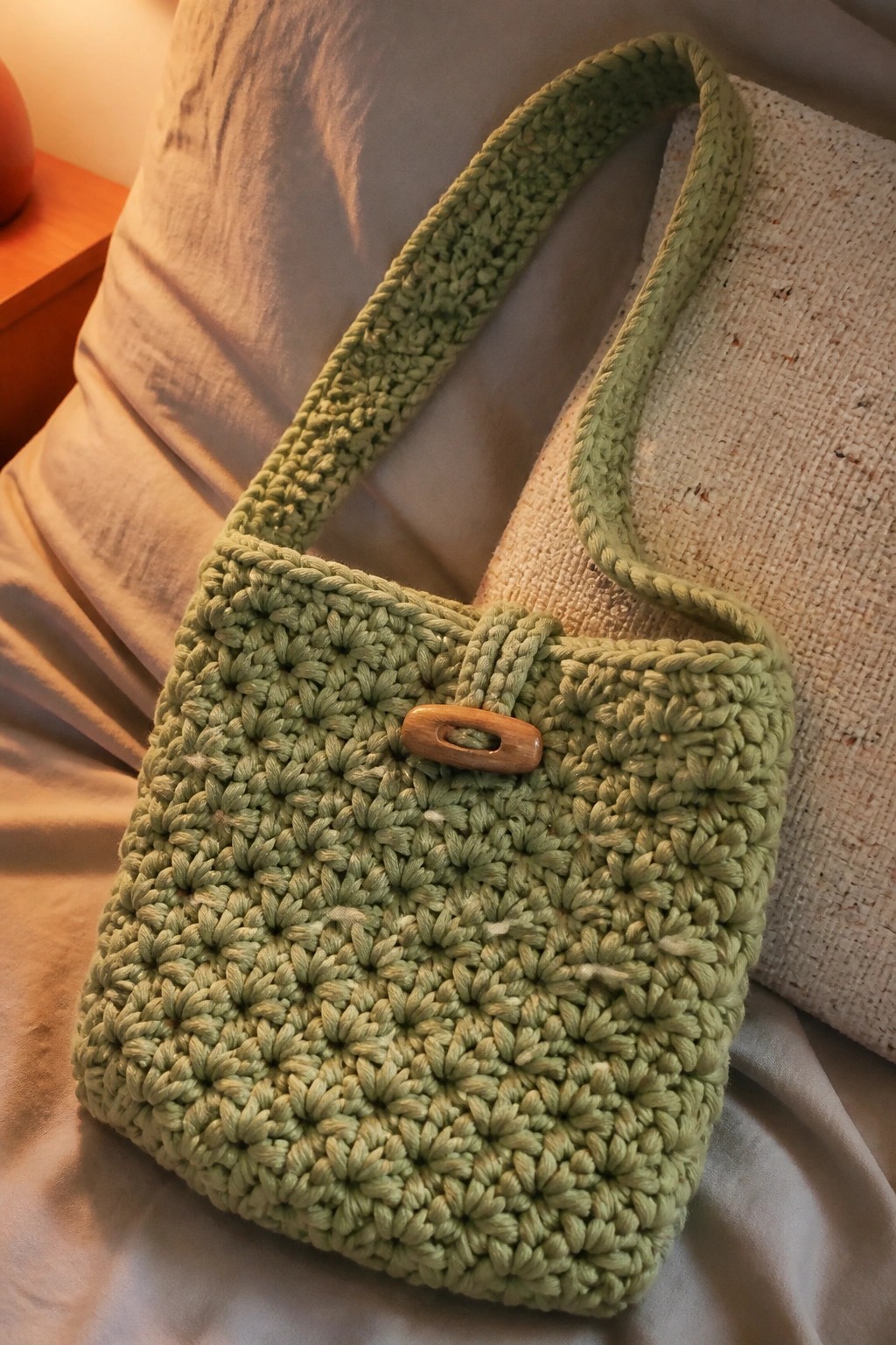 Sage green crocheted handbag with wooden button on white linen bedding