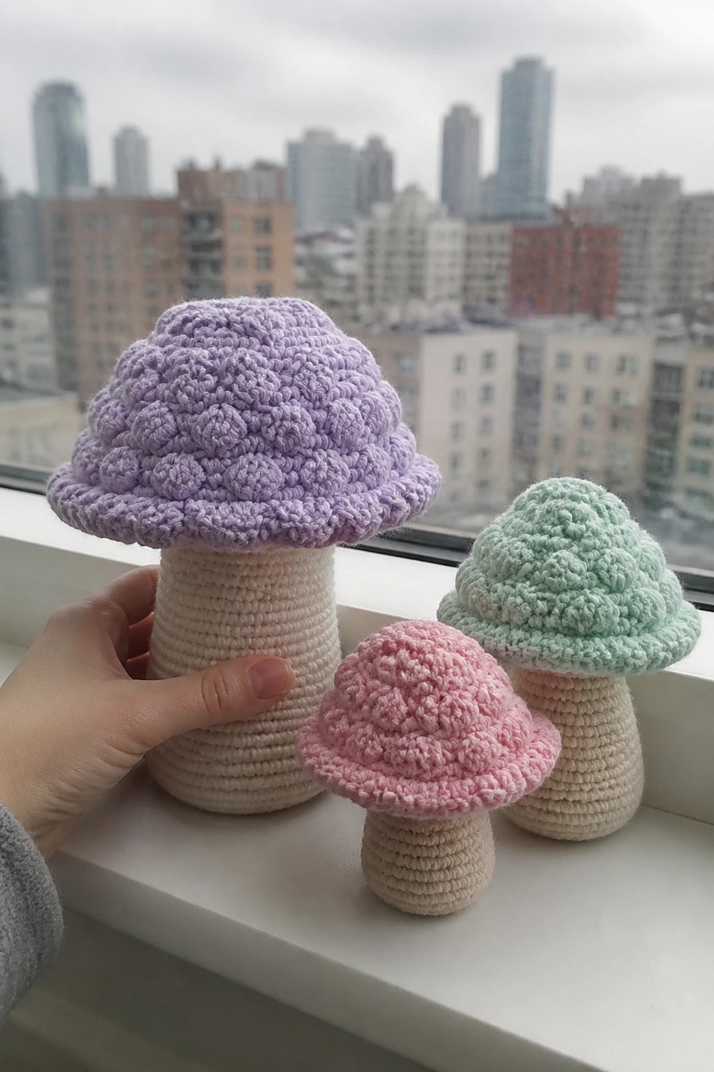 Three crocheted mushrooms with purple, pink, and mint caps on beige stems placed on a windowsill.