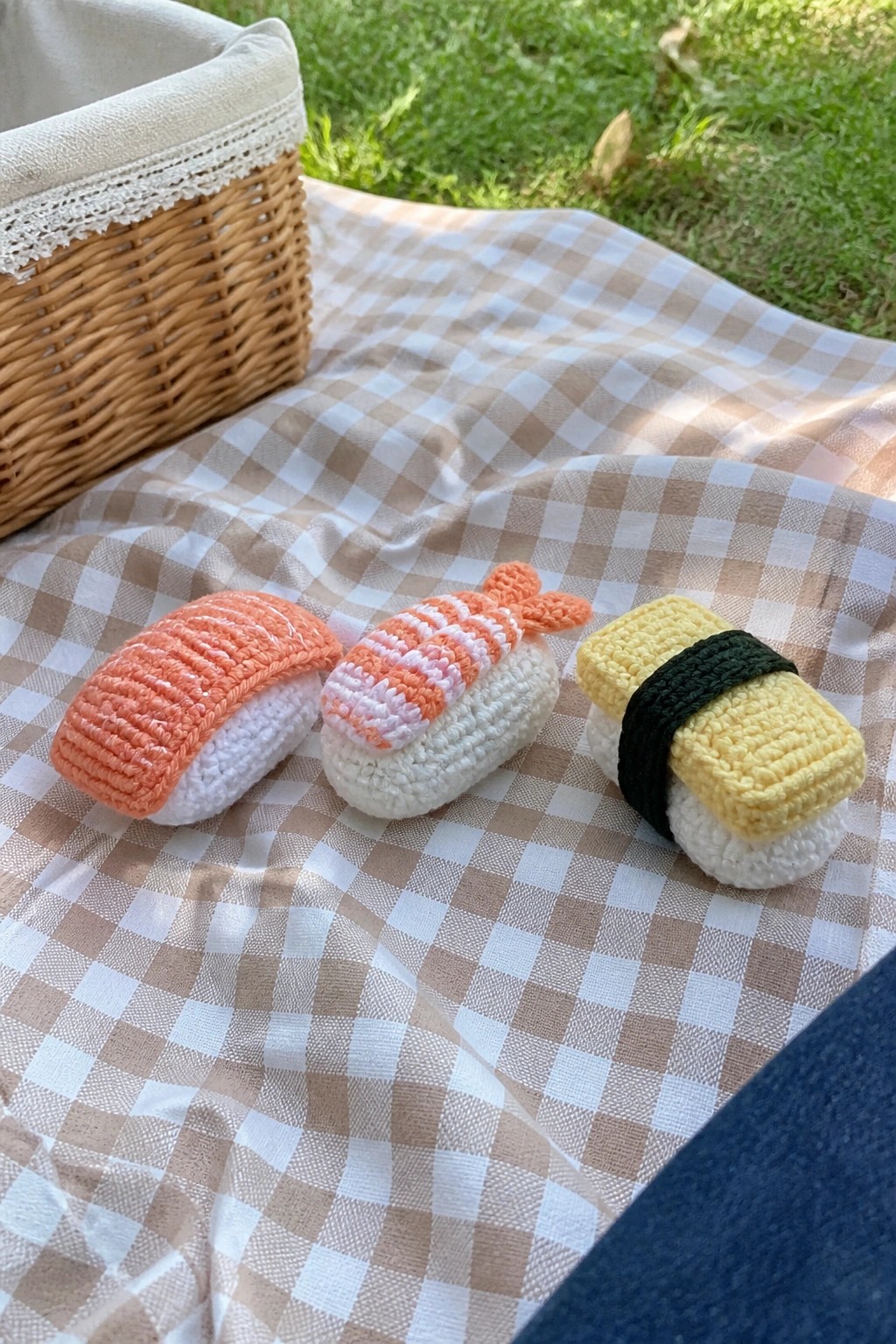 Three crocheted sushi pieces sit on a gingham blanket beside a wicker basket.