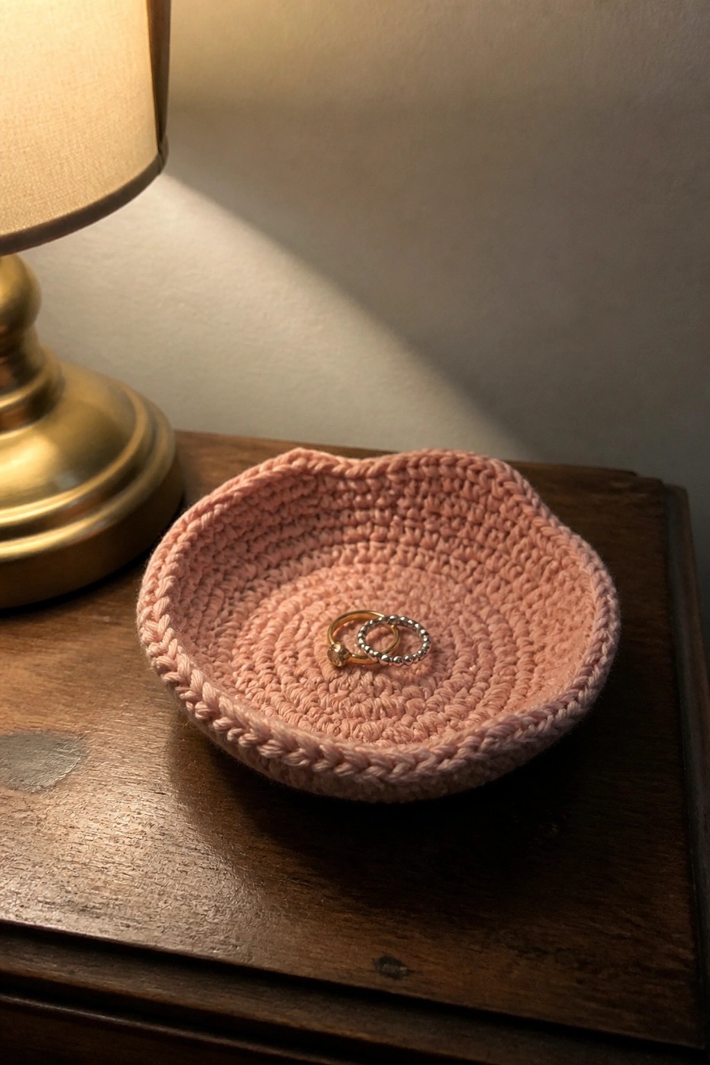 Pink crocheted bowl holding two rings on wooden table beside brass lamp base
