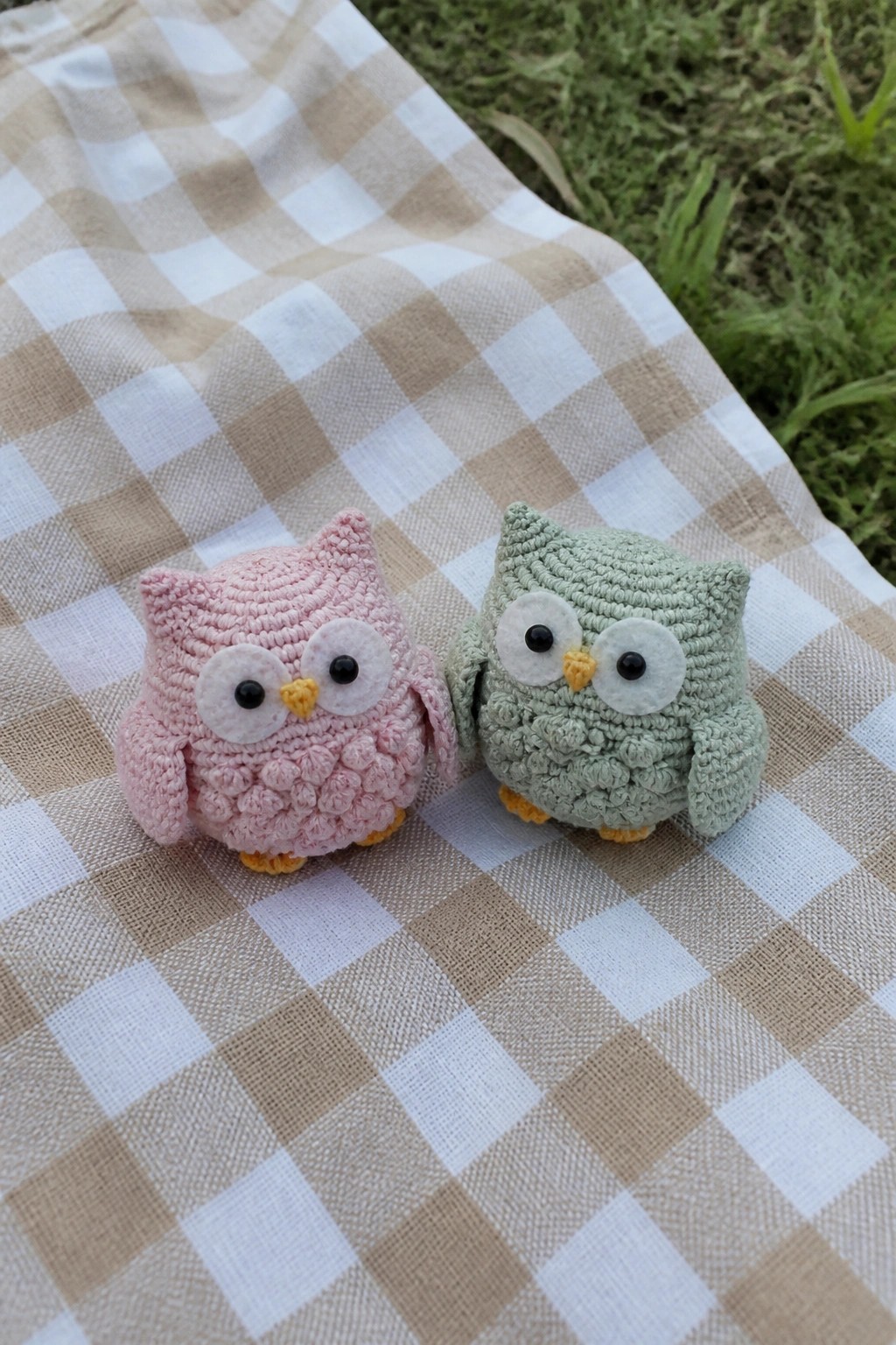 Two small crocheted owls, one pink and one green, on a checkered blanket.