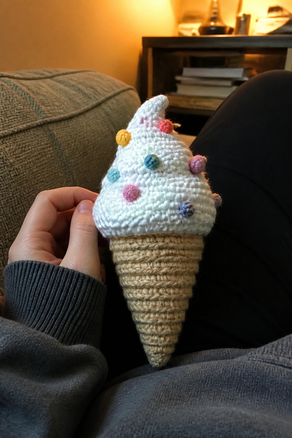 Hand holding crocheted white ice cream cone with colorful pom-poms on beige base.