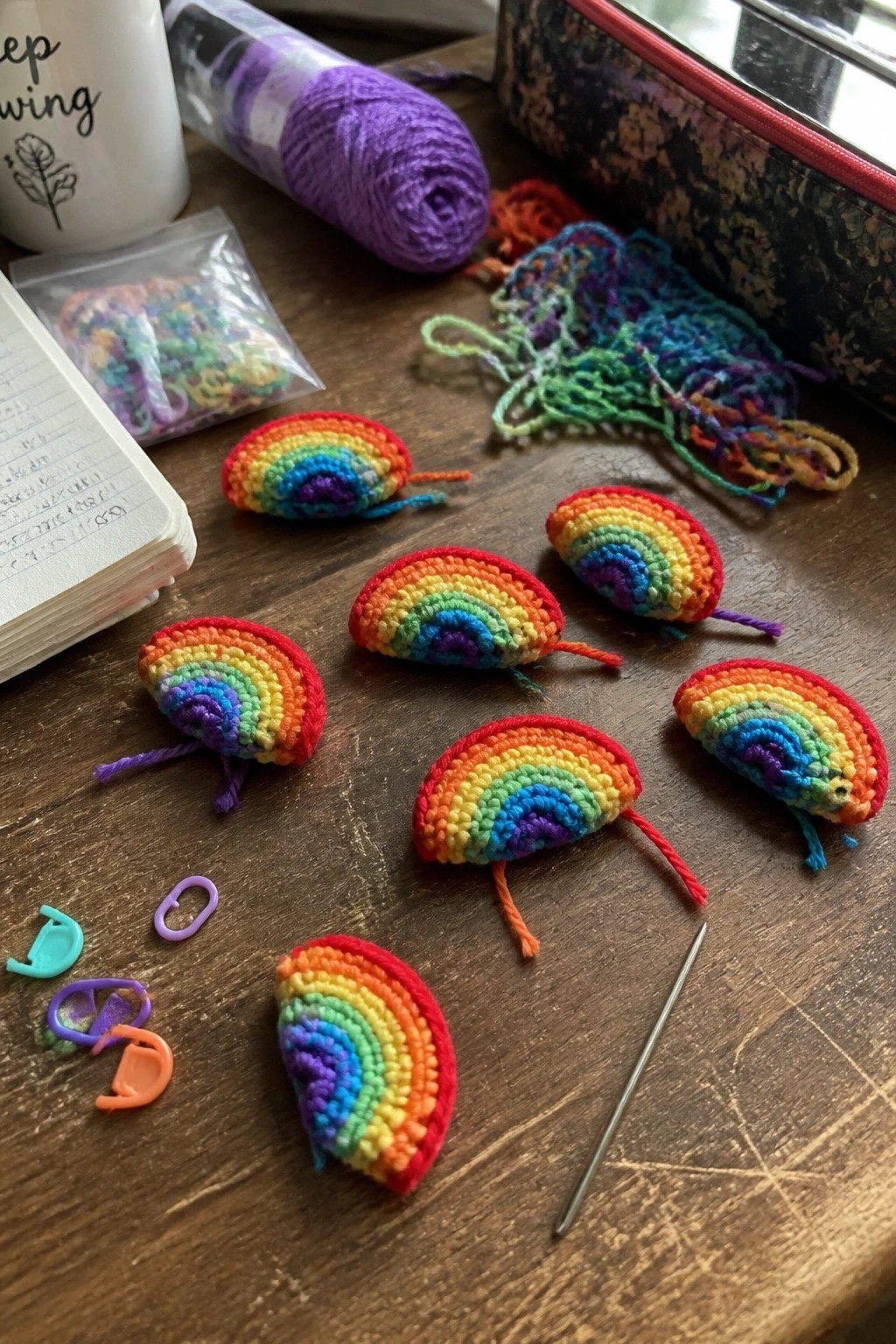 Crocheted rainbow semicircles with legs amid yarn, markers, and notebook on table