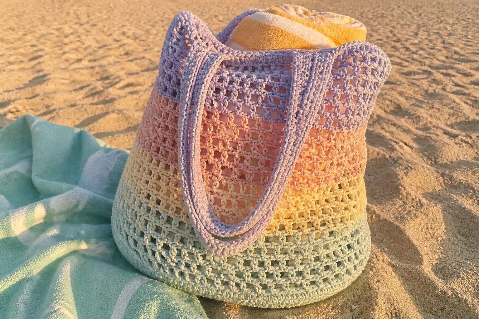 Rainbow crocheted mesh tote bag on beach sand beside turquoise towel.