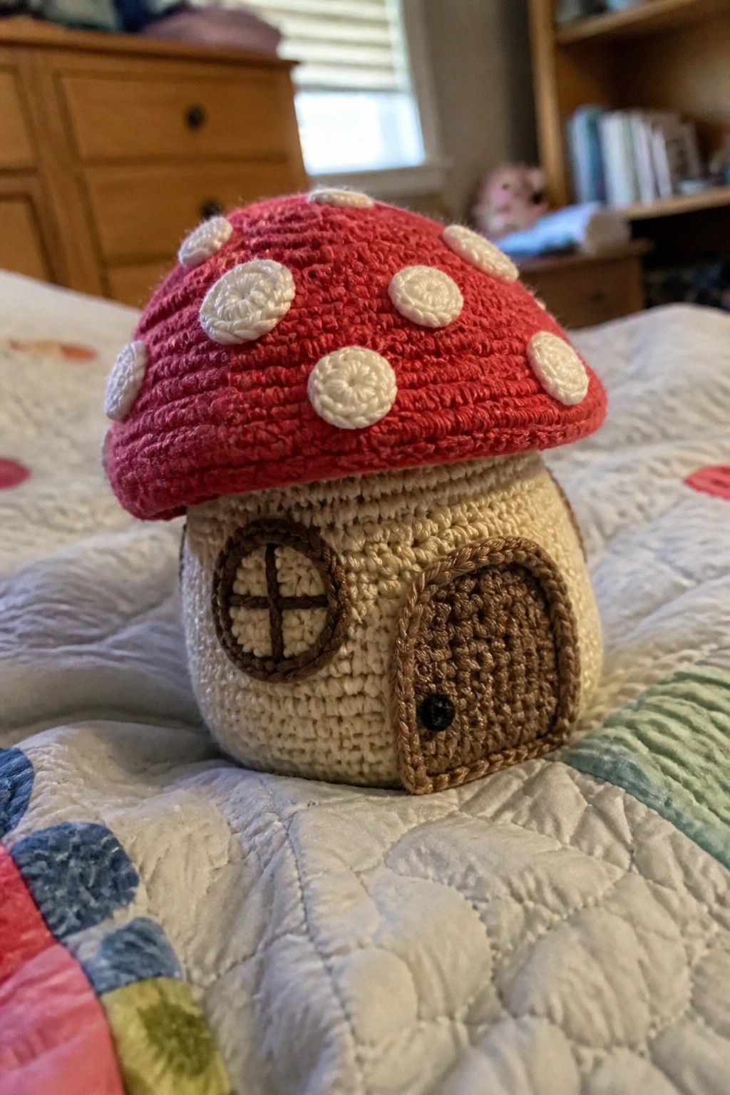 Crocheted mushroom house with red spotted cap, brown door, and window on quilt