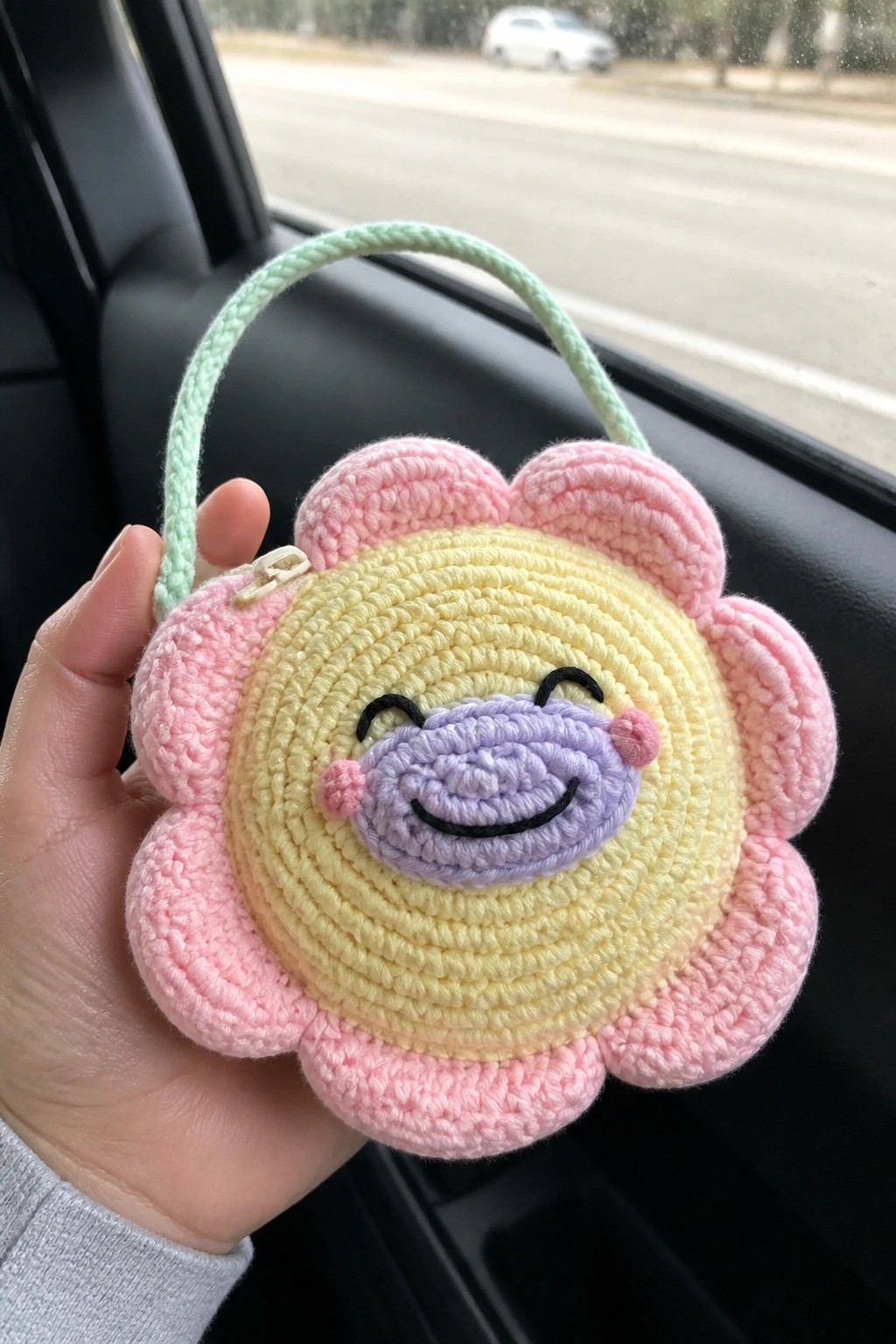 Hand holding pink crocheted flower pouch with yellow center, smiling face, and green strap