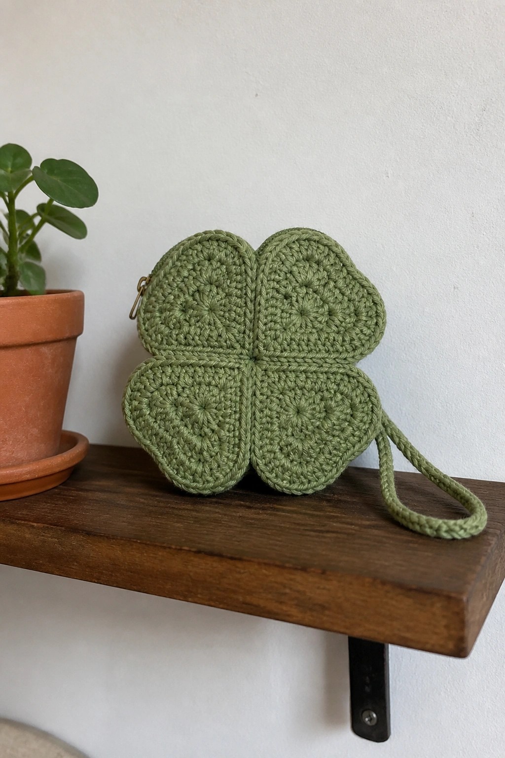 Green crocheted four-leaf clover pouch with strap on wooden shelf beside potted plant