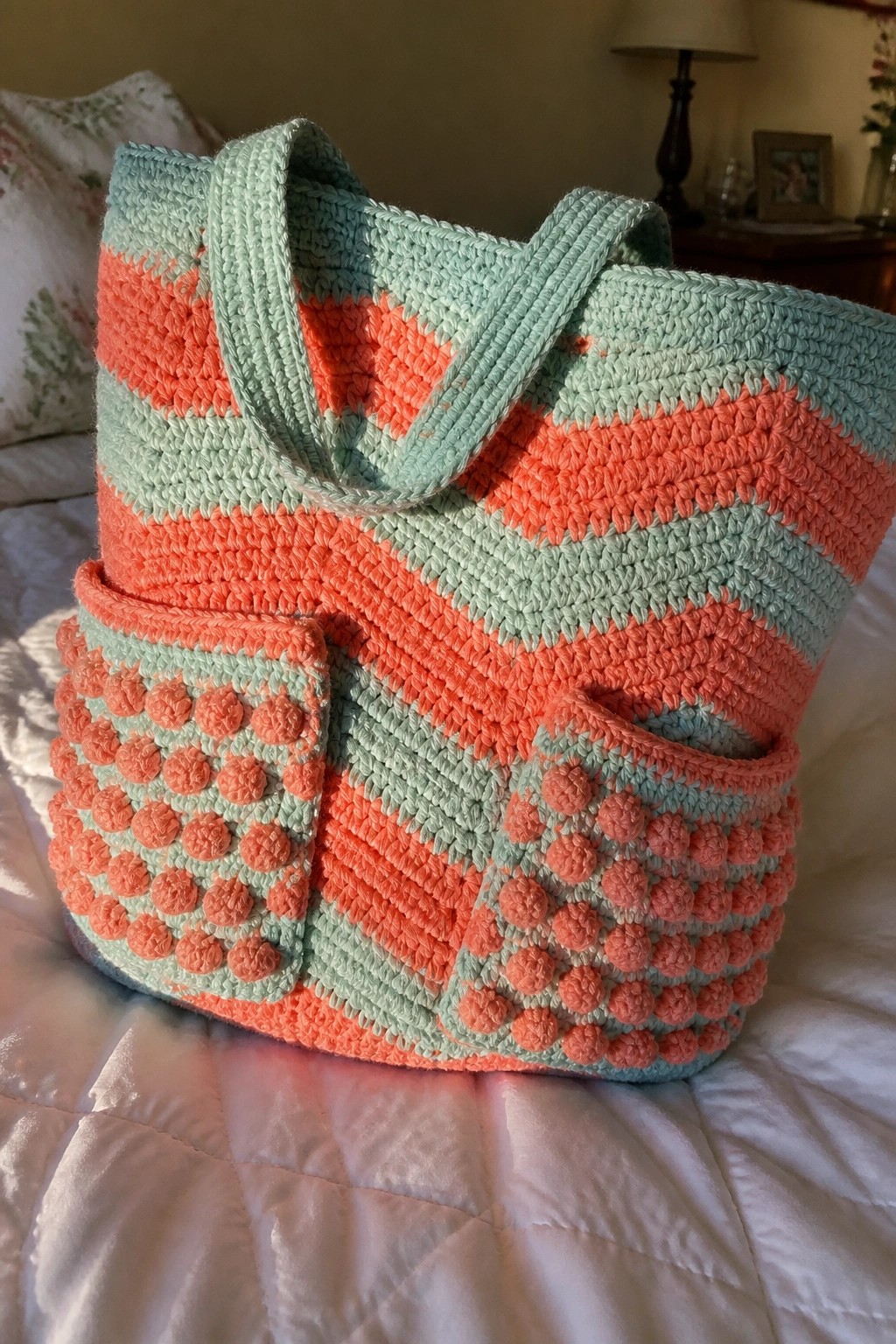 Crocheted tote bag with coral and mint stripes, hearts, and bobble pockets on bed