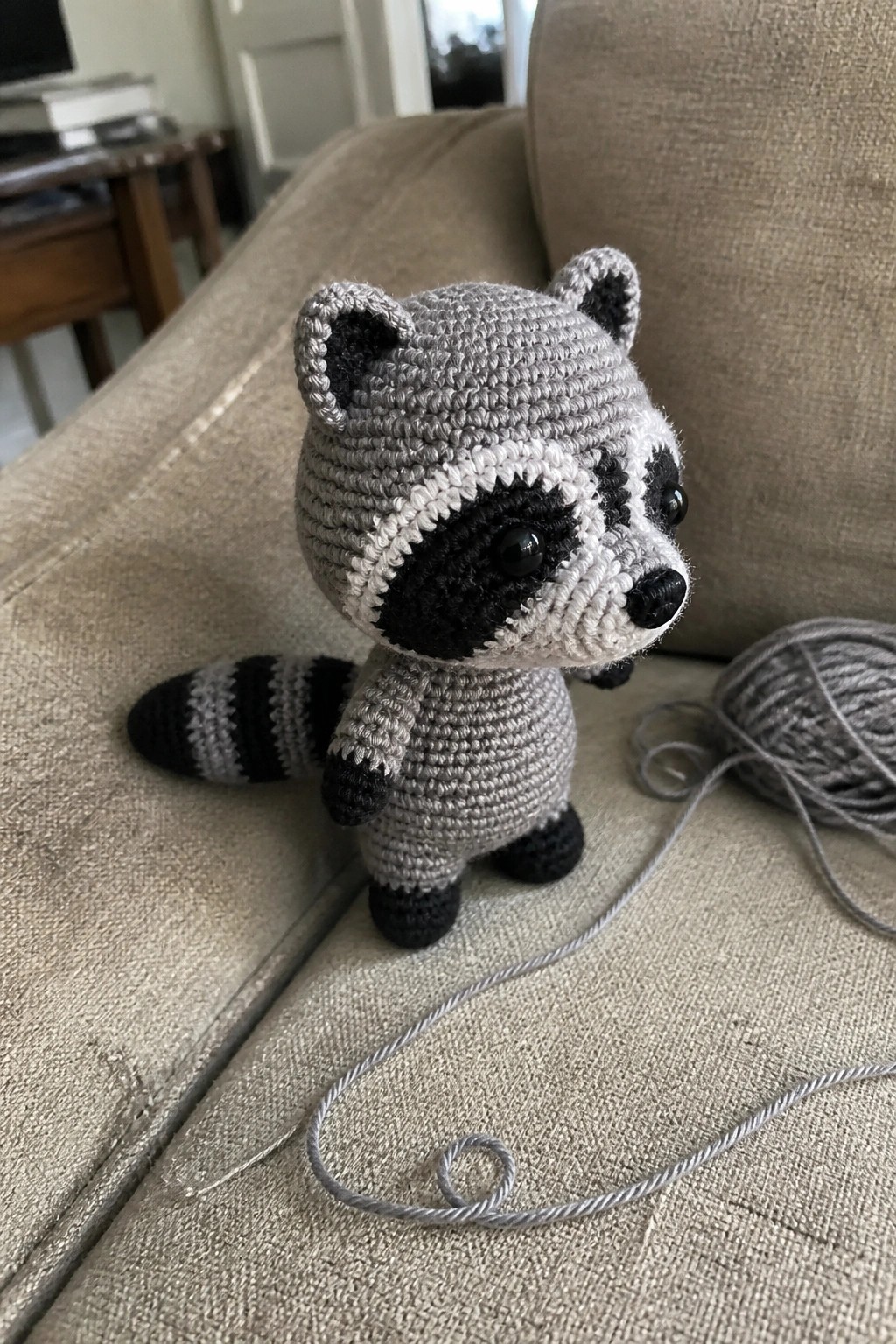 Crocheted gray raccoon toy on beige couch beside ball of yarn