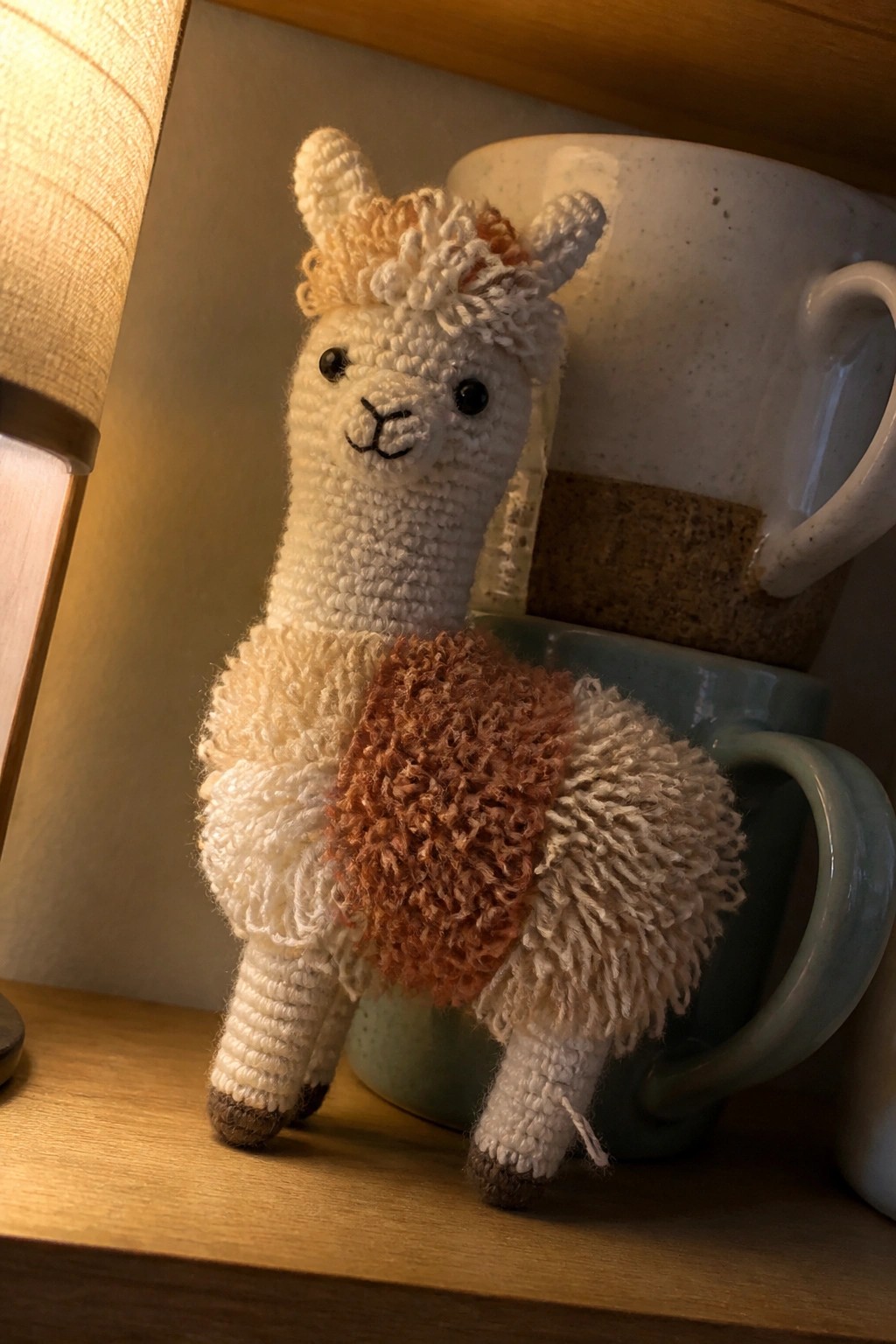 Crocheted white llama with fluffy brown and beige patches on wooden shelf beside mugs.