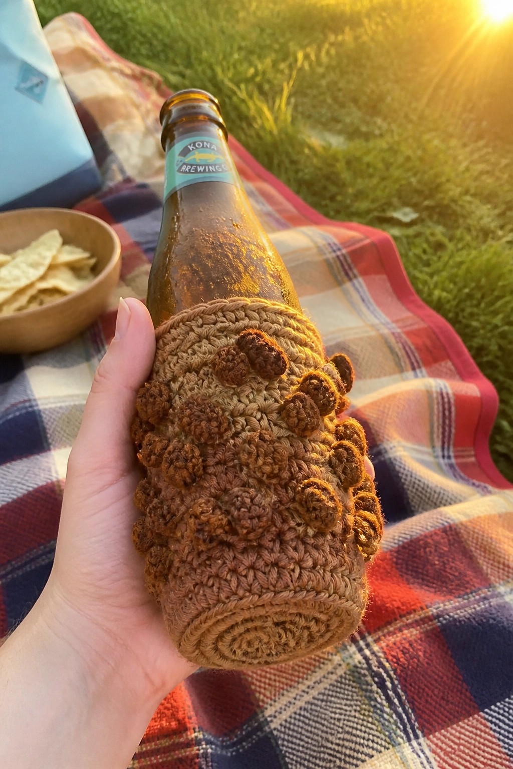 Hand holds Kona beer bottle in bumpy brown crocheted cozy on plaid blanket