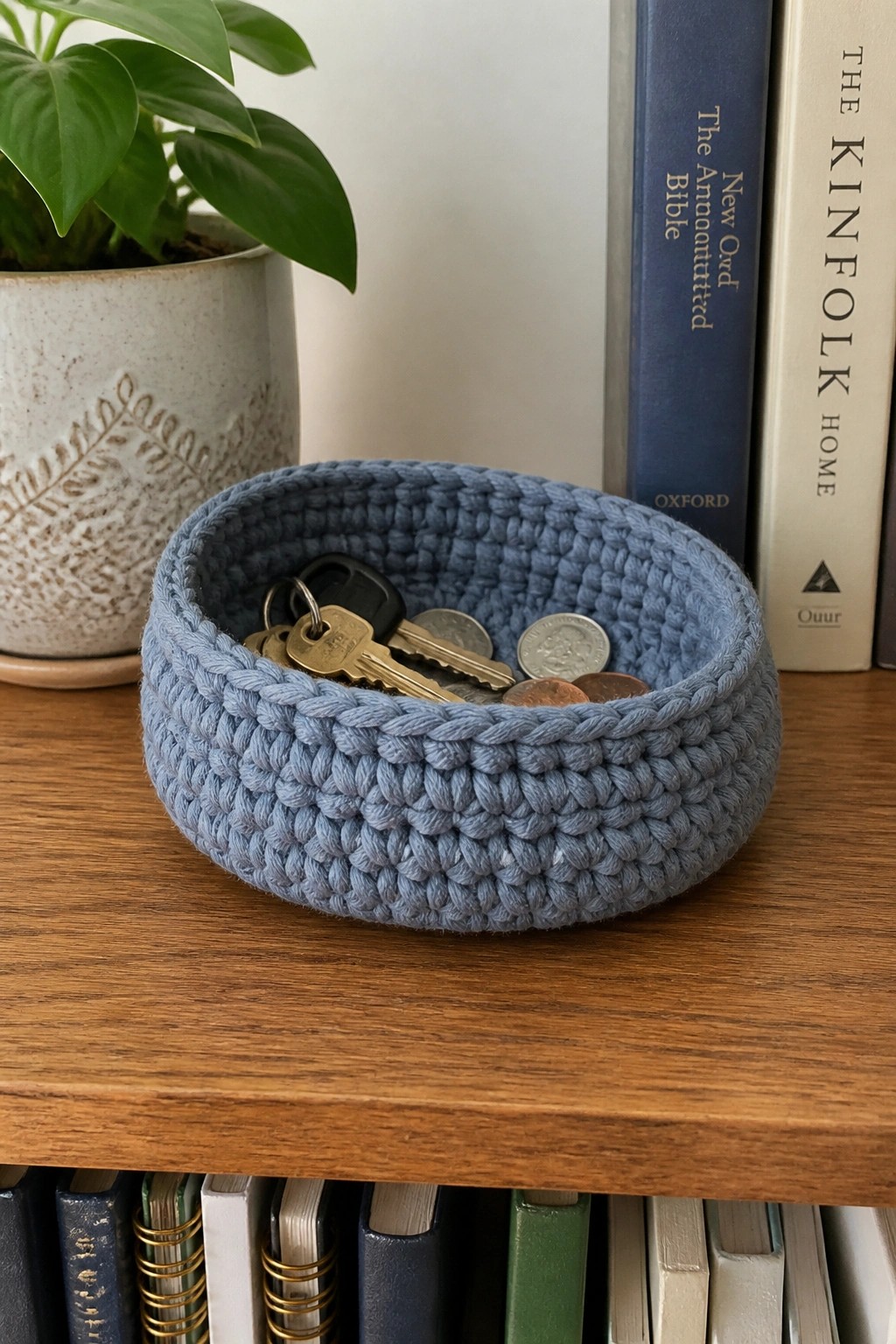 Blue crocheted bowl with keys and coins on wooden shelf near books and plant