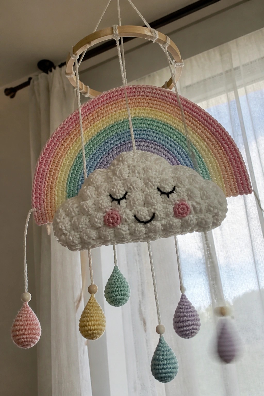 Crocheted pastel rainbow arch over smiling cloud with hanging yarn raindrops on hoop.