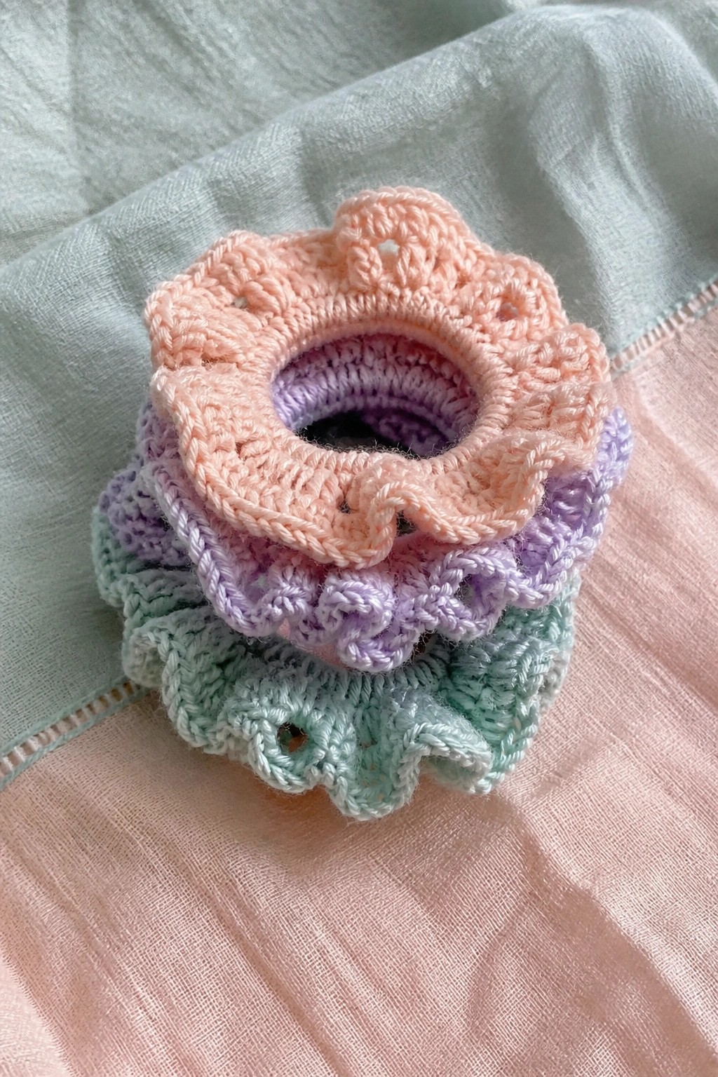 Pastel crocheted scrunchies in peach, purple, and mint stacked on fabric.