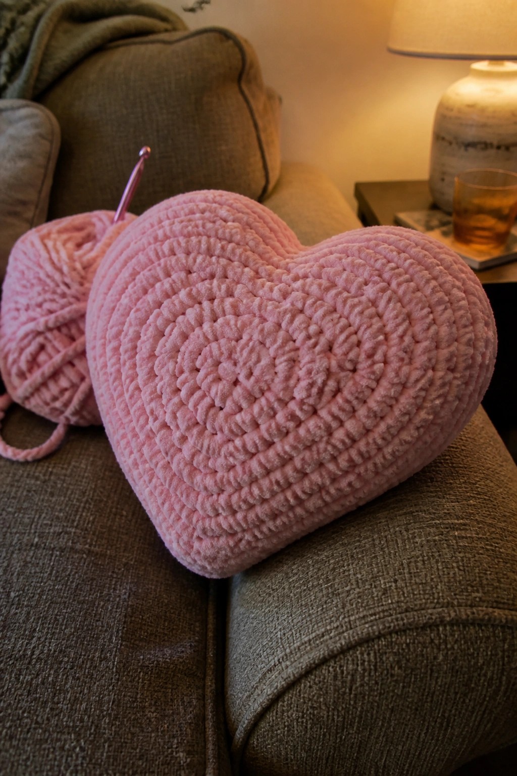 Pink crocheted heart pillow on gray couch beside yarn ball and hook