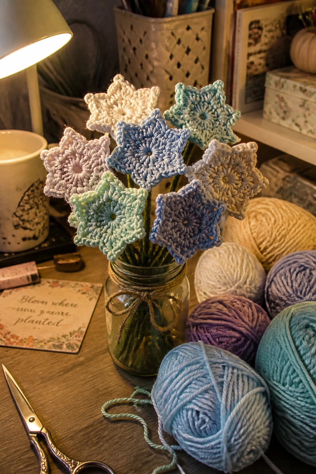 Crocheted star-shaped flowers in pastel colors arranged in a glass jar with yarn balls nearby on a wooden table.