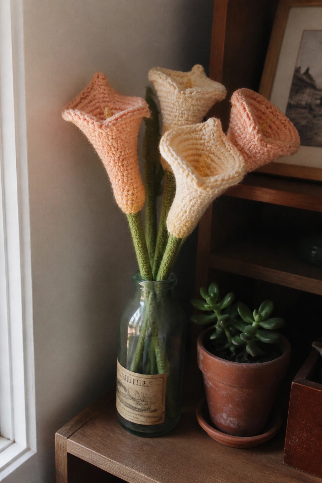 Crocheted peach and cream calla lilies with green stems in a glass bottle beside succulent