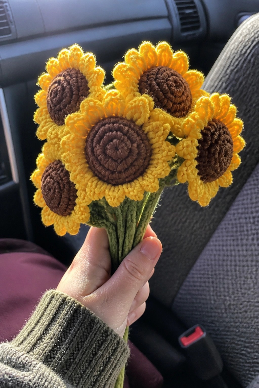 Hand in green sweater holding yellow-brown crocheted sunflower bouquet inside a car