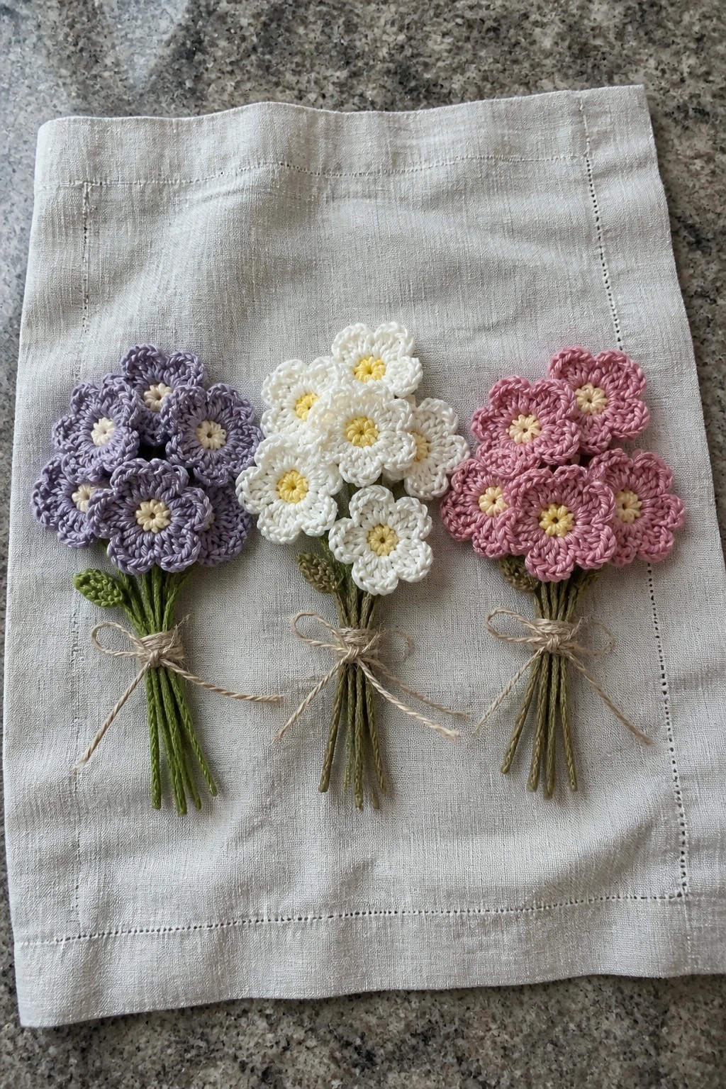Three small crocheted flower bouquets in purple, white, and pink sit tied with twine on a linen cloth.