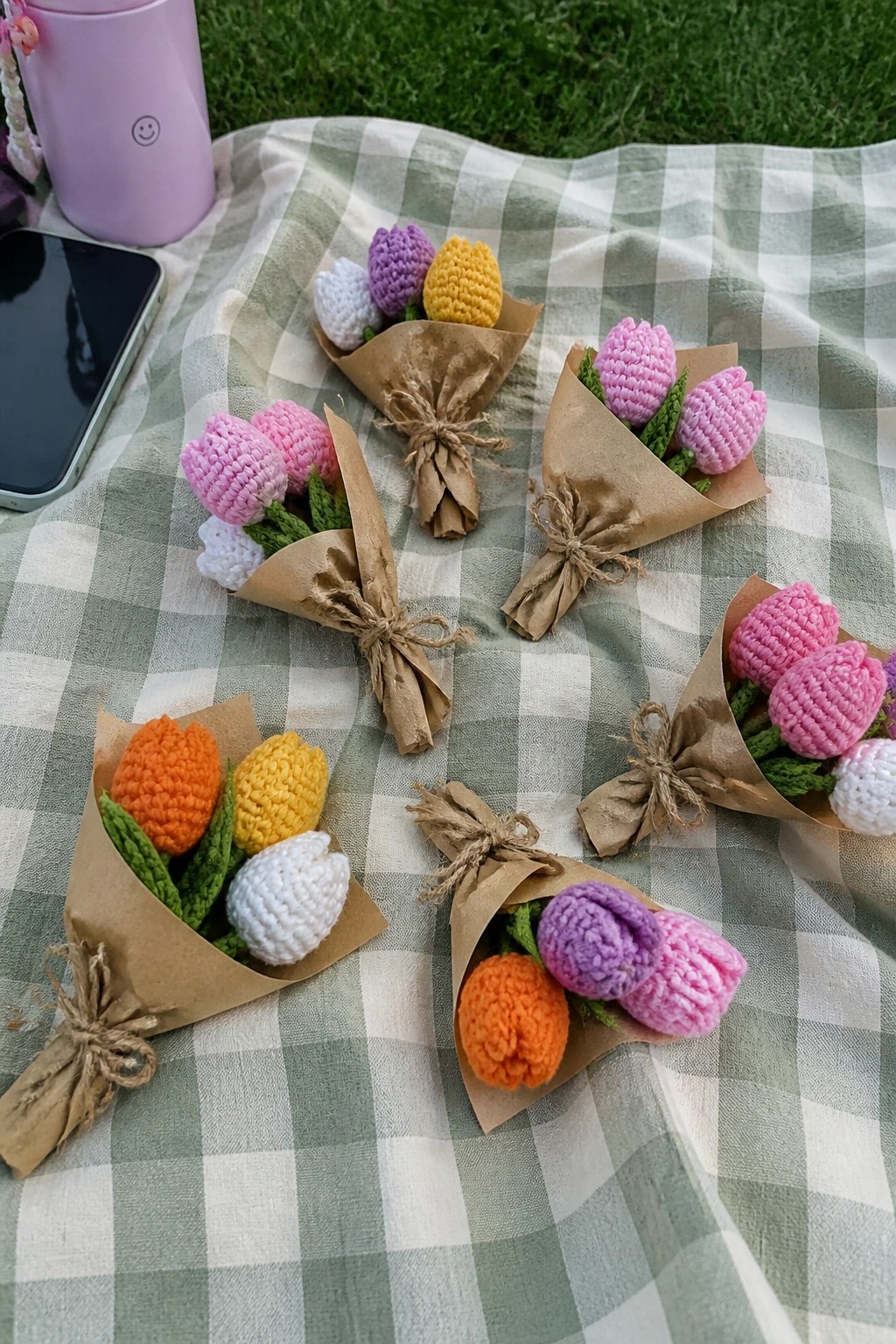 Small crocheted tulip bouquets in pink, purple, yellow, orange, and white wrapped in brown paper and tied with twine on a picnic blanket.