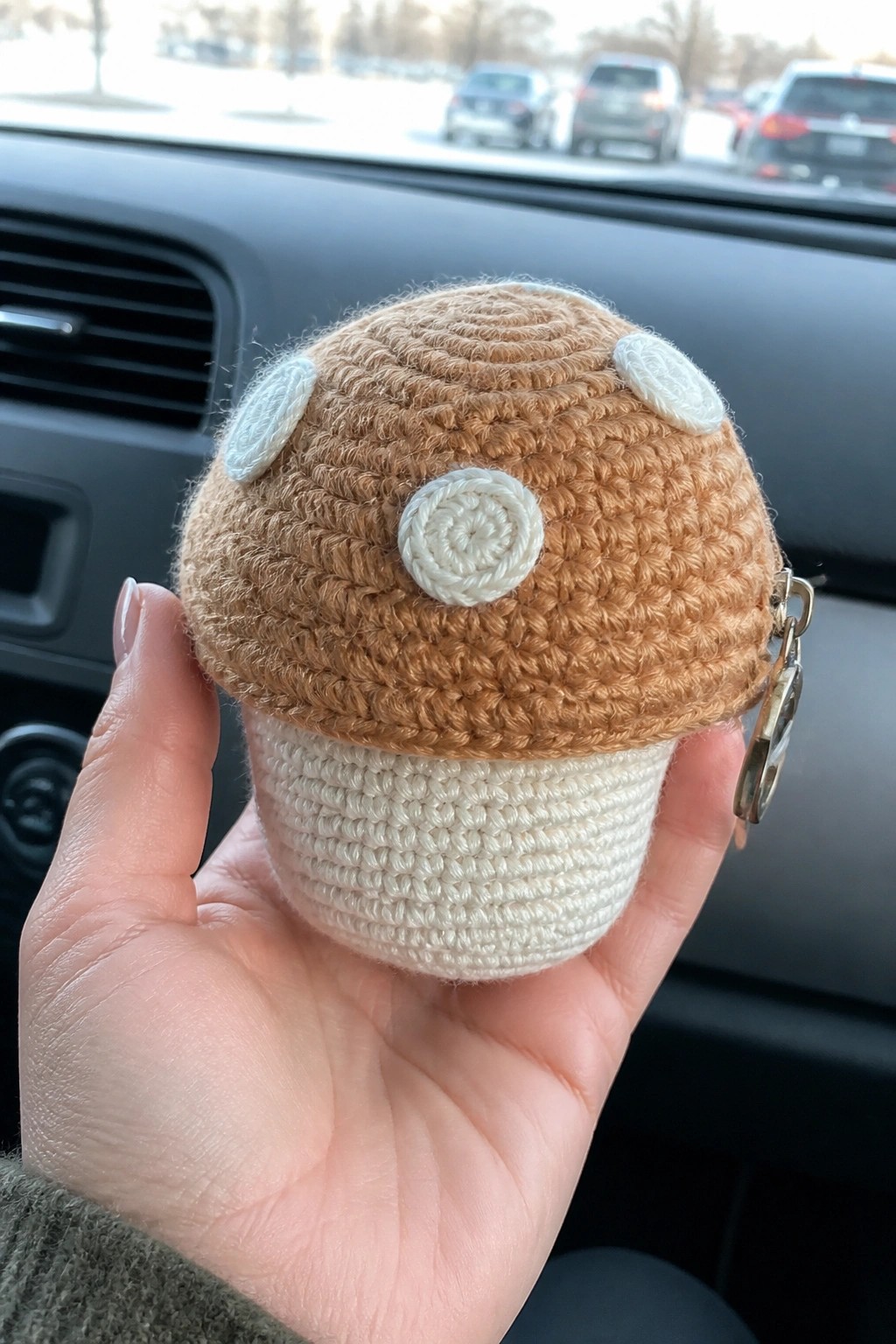 Hand holds crocheted brown mushroom keychain with white spots inside car