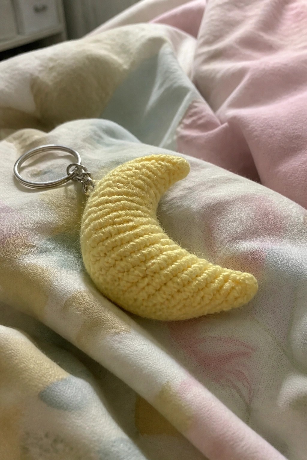 Yellow crocheted crescent moon keychain on pastel floral patterned blanket.