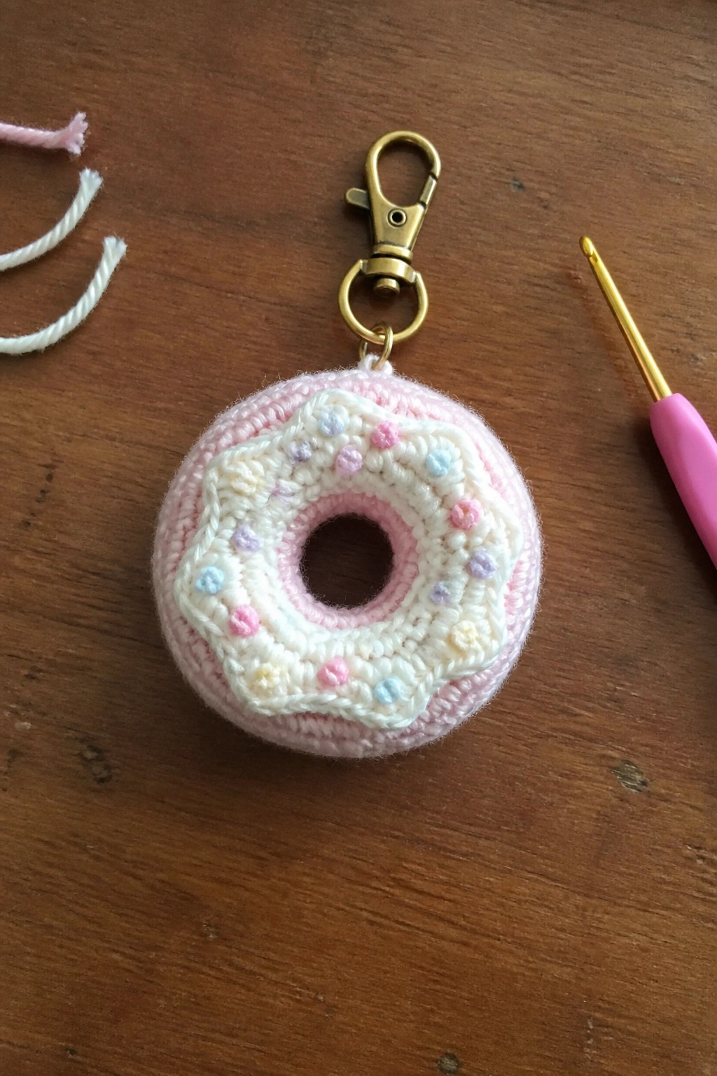 Crocheted pink donut keychain with sprinkles, gold clasp, and pink hook on wood.