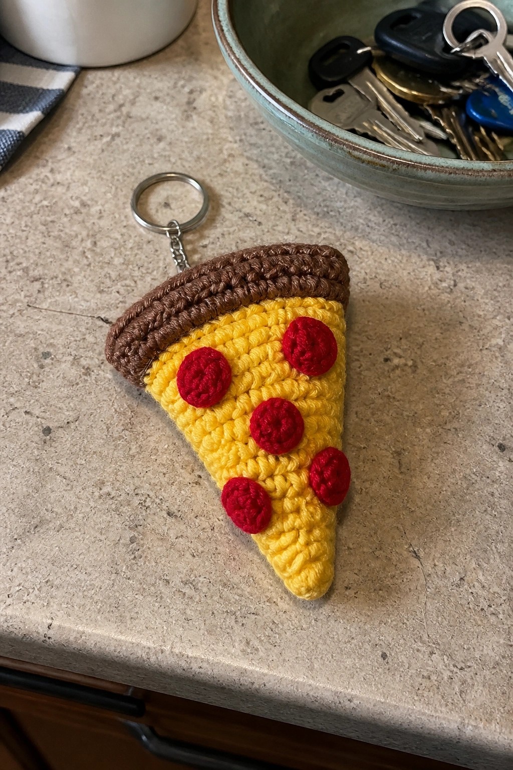 Crocheted yellow pizza slice keychain with red pepperoni and brown crust on countertop