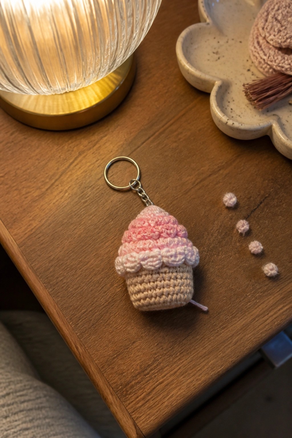 Crocheted pink cupcake keychain on wooden table near lit lamp and dish