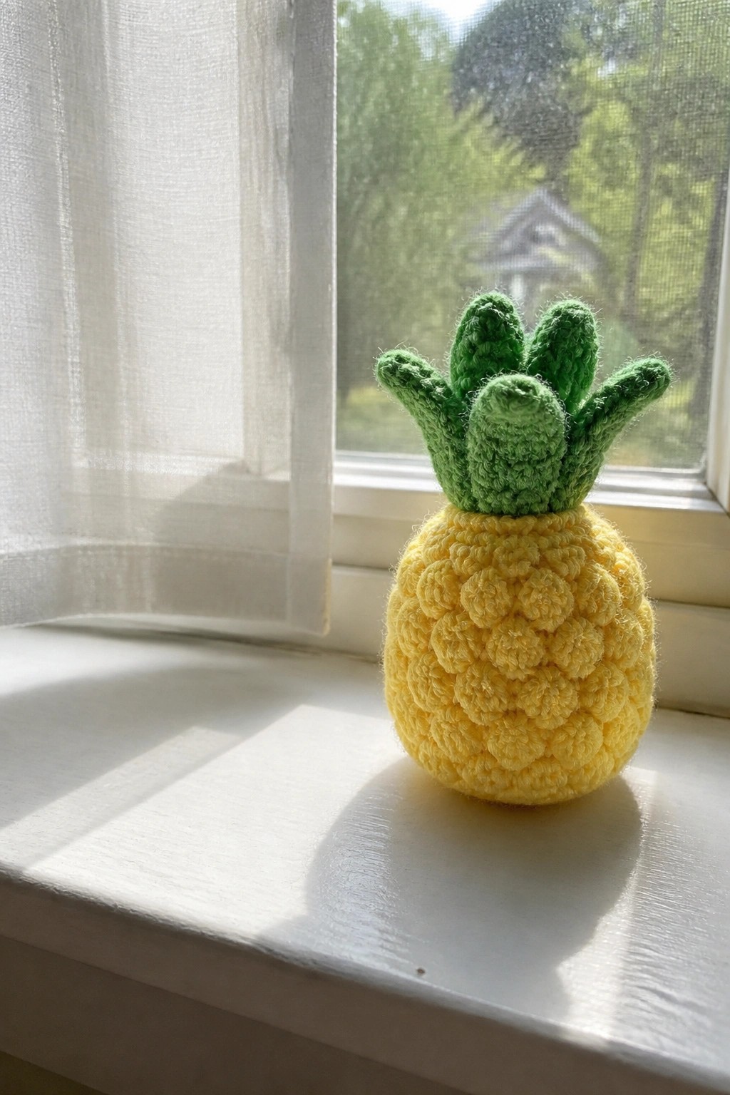 Yellow crocheted pineapple with green leaves on sunny white windowsill by window.