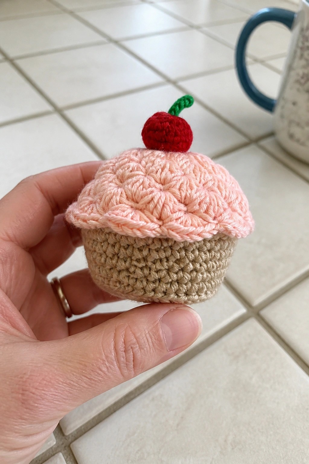 Hand holding crocheted cupcake with pink top, beige base, and red cherry