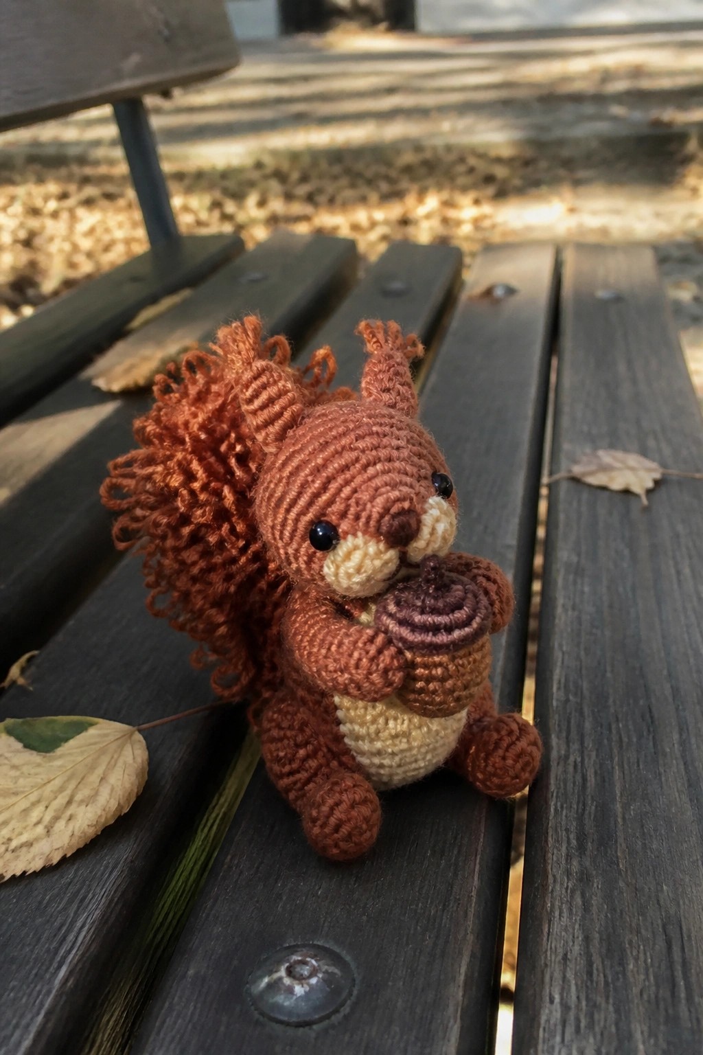 A small brown crocheted squirrel holding an acorn sits on a wooden bench.