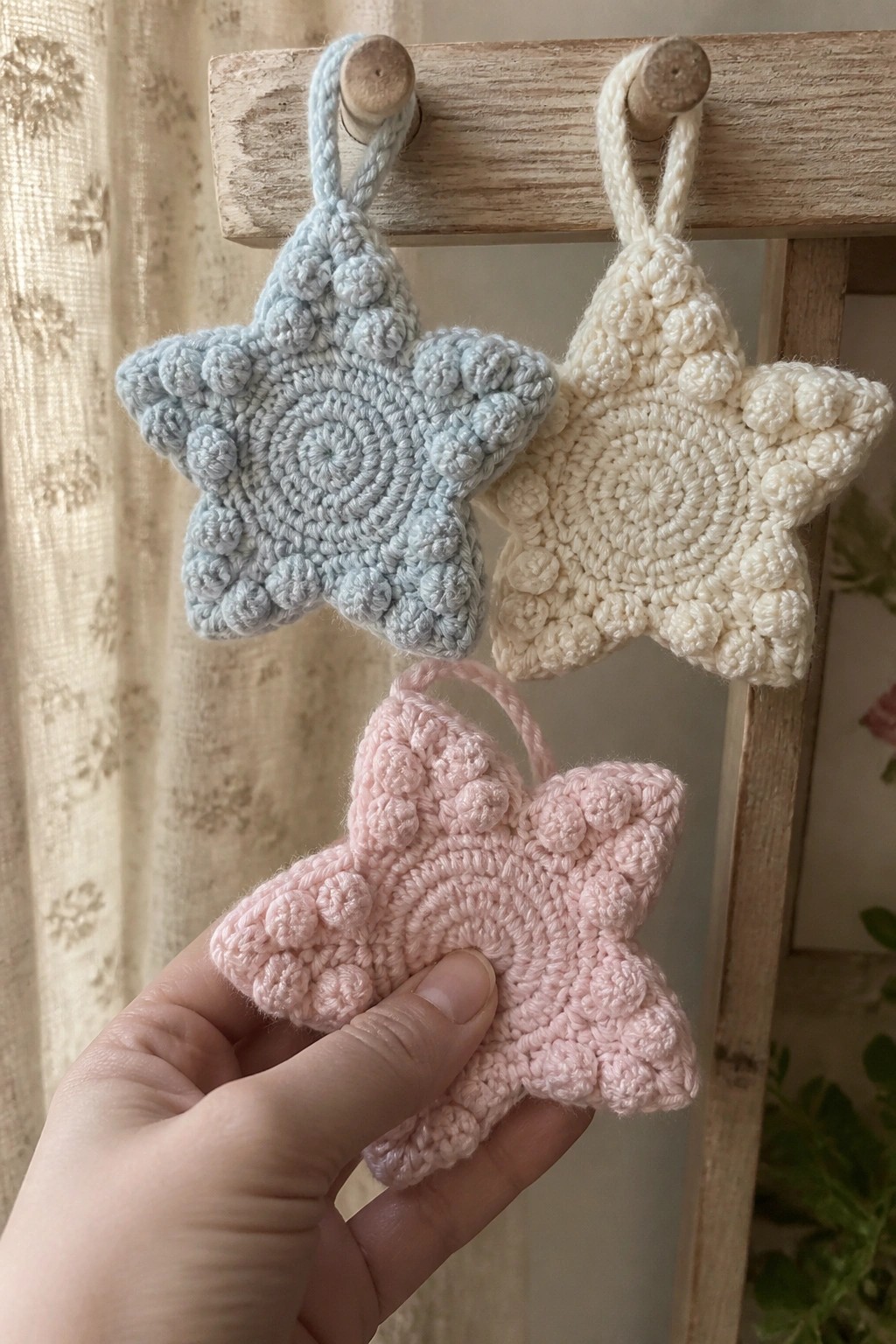 Three pastel crocheted stars in blue, cream, and pink hanging on wooden pegs.