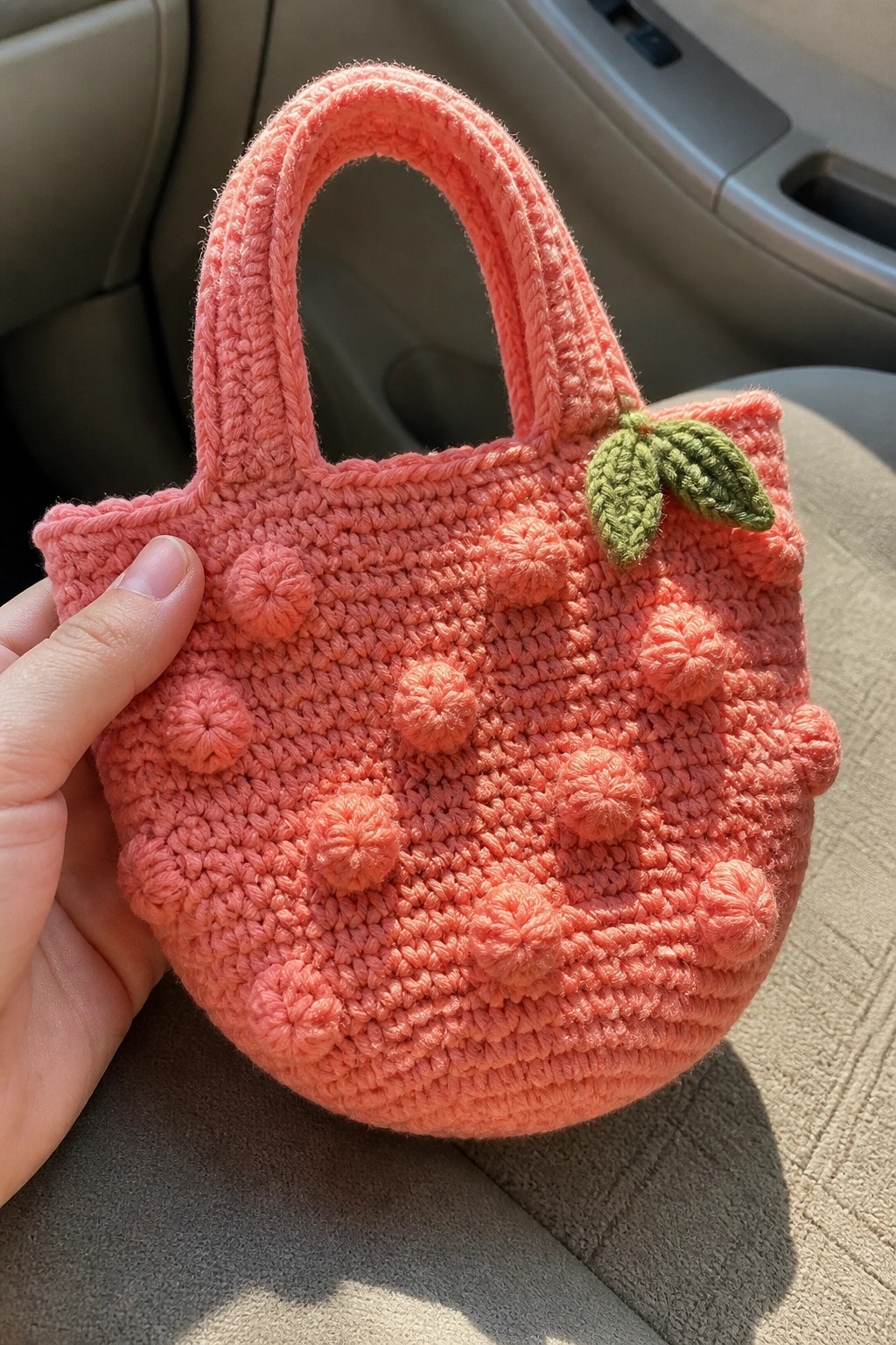 Hand holding pink crocheted round bag with bobble texture and green leaves in car